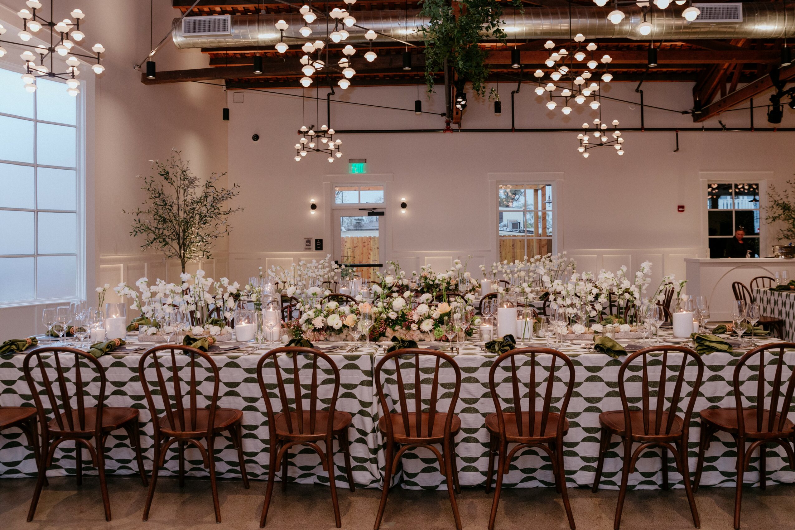 Long banquet table with floral centerpieces, surrounded by wooden chairs, in a decorated event space with hanging lights.