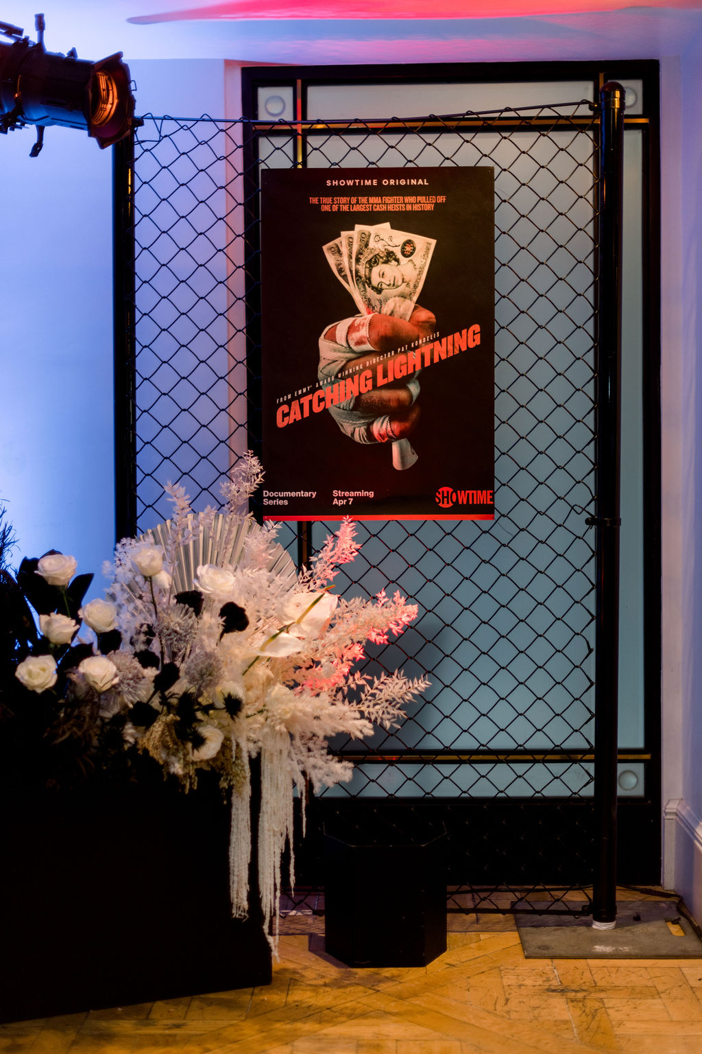 Poster on a metal grid display features a hand holding cash with the title 'CATCHING LIGHTNING' in red, surrounded by flowers.