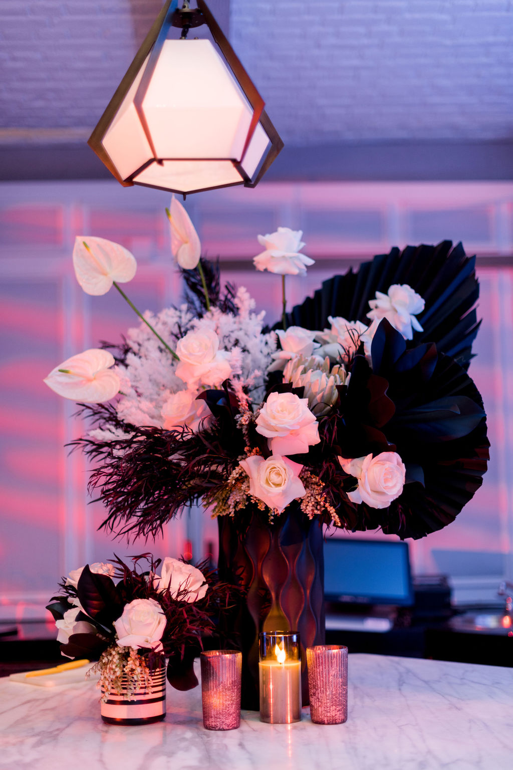 A floral arrangement with white flowers and dark leaves, illuminated by candles and hanging light, on a table.
