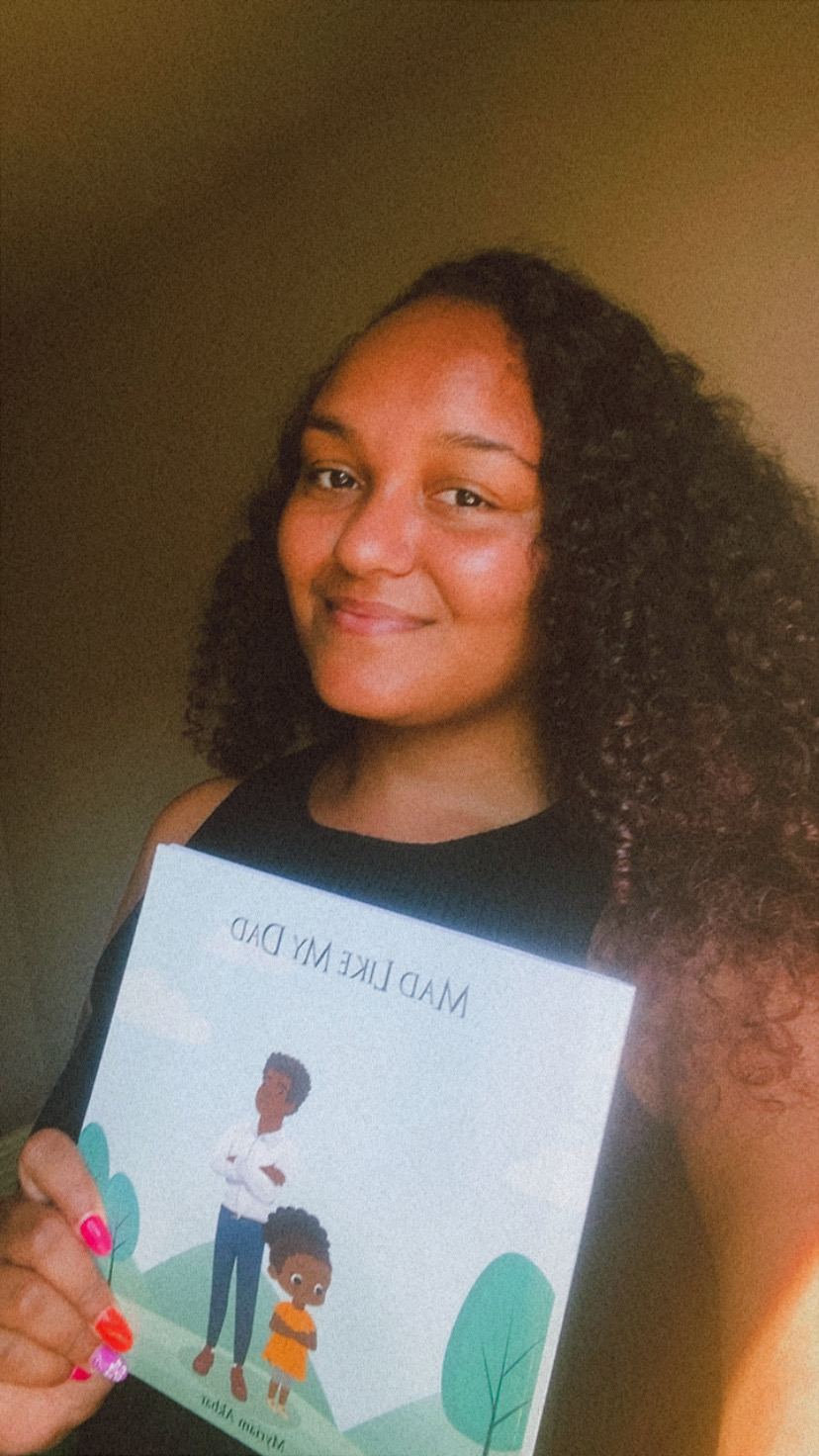 Person with curly hair holding a book or magazine with an illustration of two children and trees, smiling.