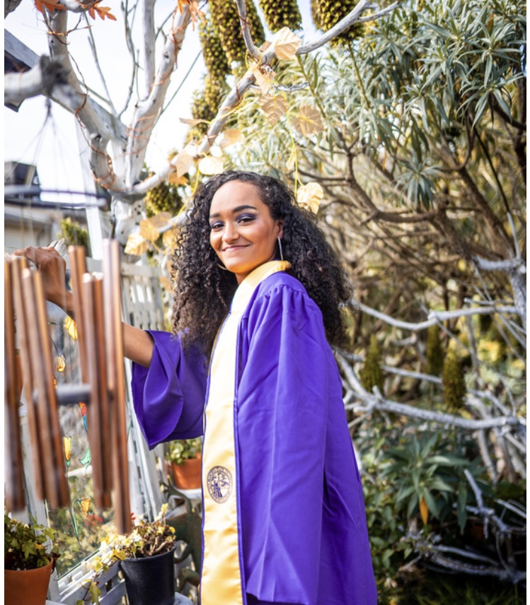 Young woman in a purple graduation gown outdoors, smiling, surrounded by trees and plants.