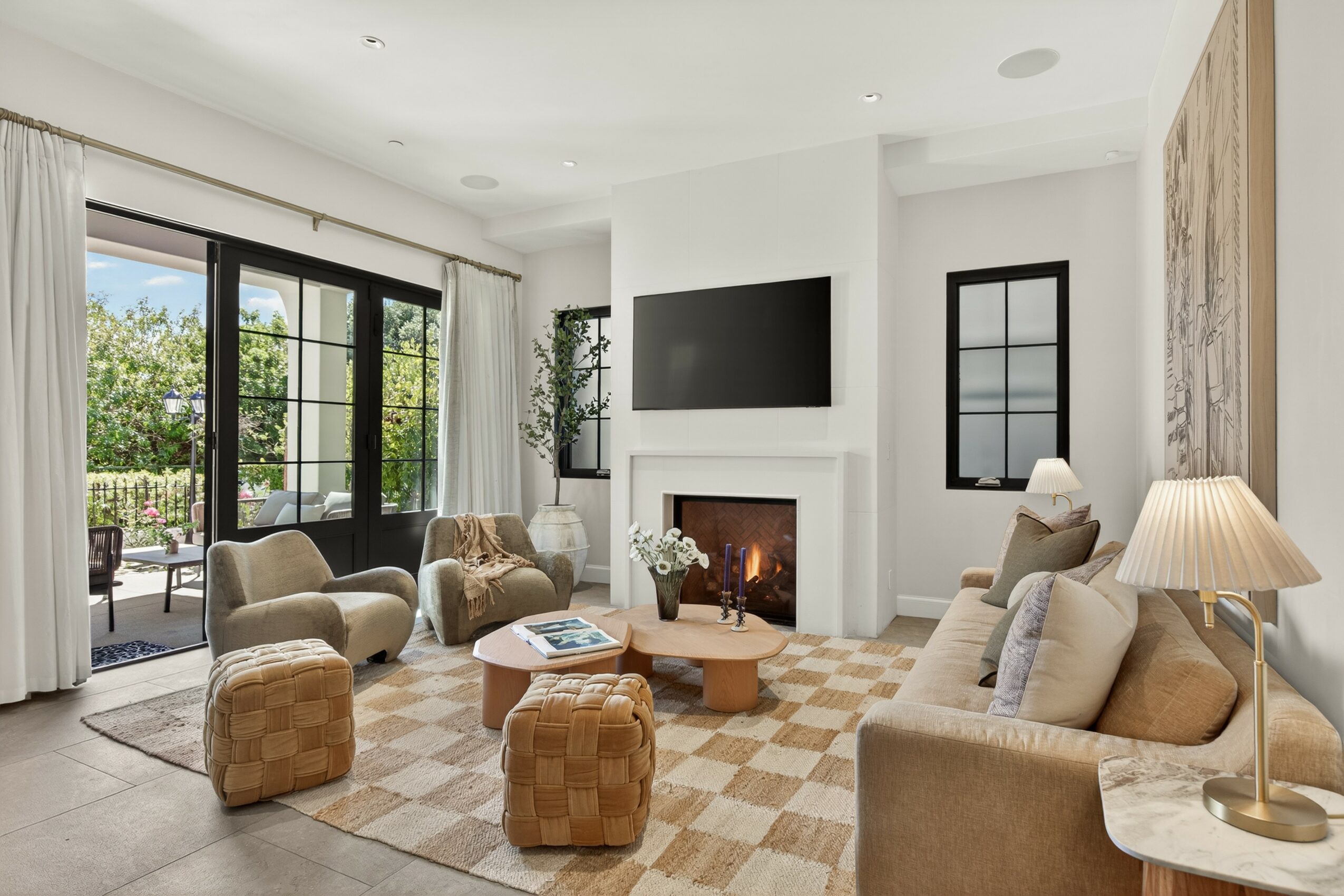 Living room with large windows, beige sofas, a fireplace, and a wall-mounted TV, decorated with plants and a side table with a lamp.