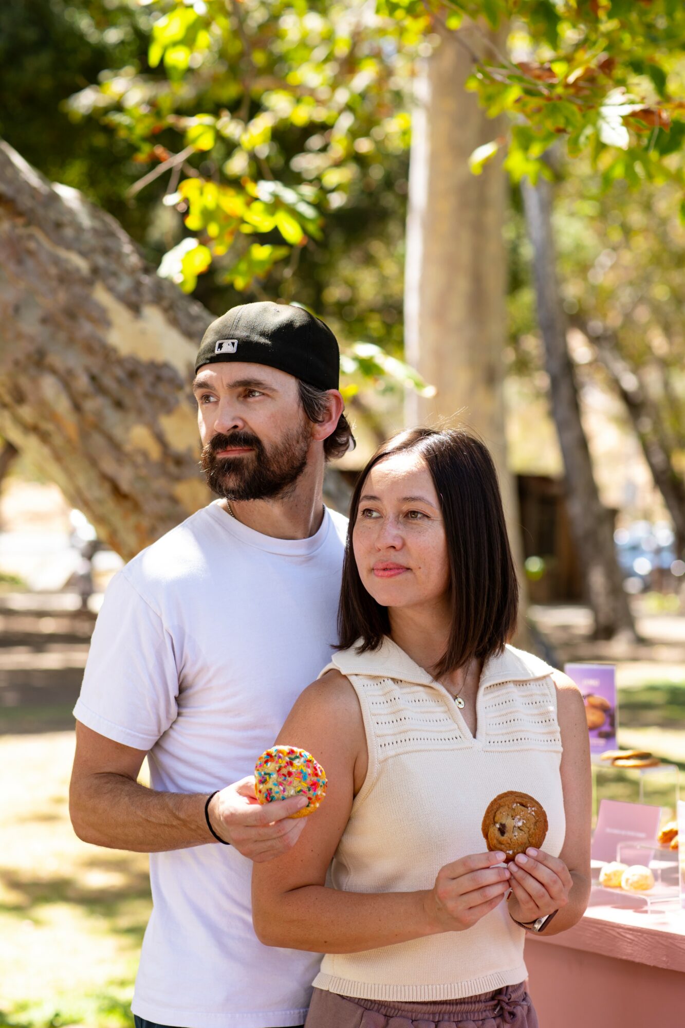A man and woman outdoors holding decorated cookies, trees and sunlight in background.