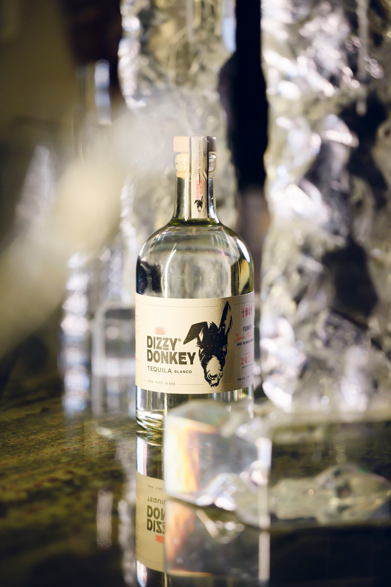A clear glass bottle with a label featuring a donkey silhouette, placed on a reflective surface with a blurred background.