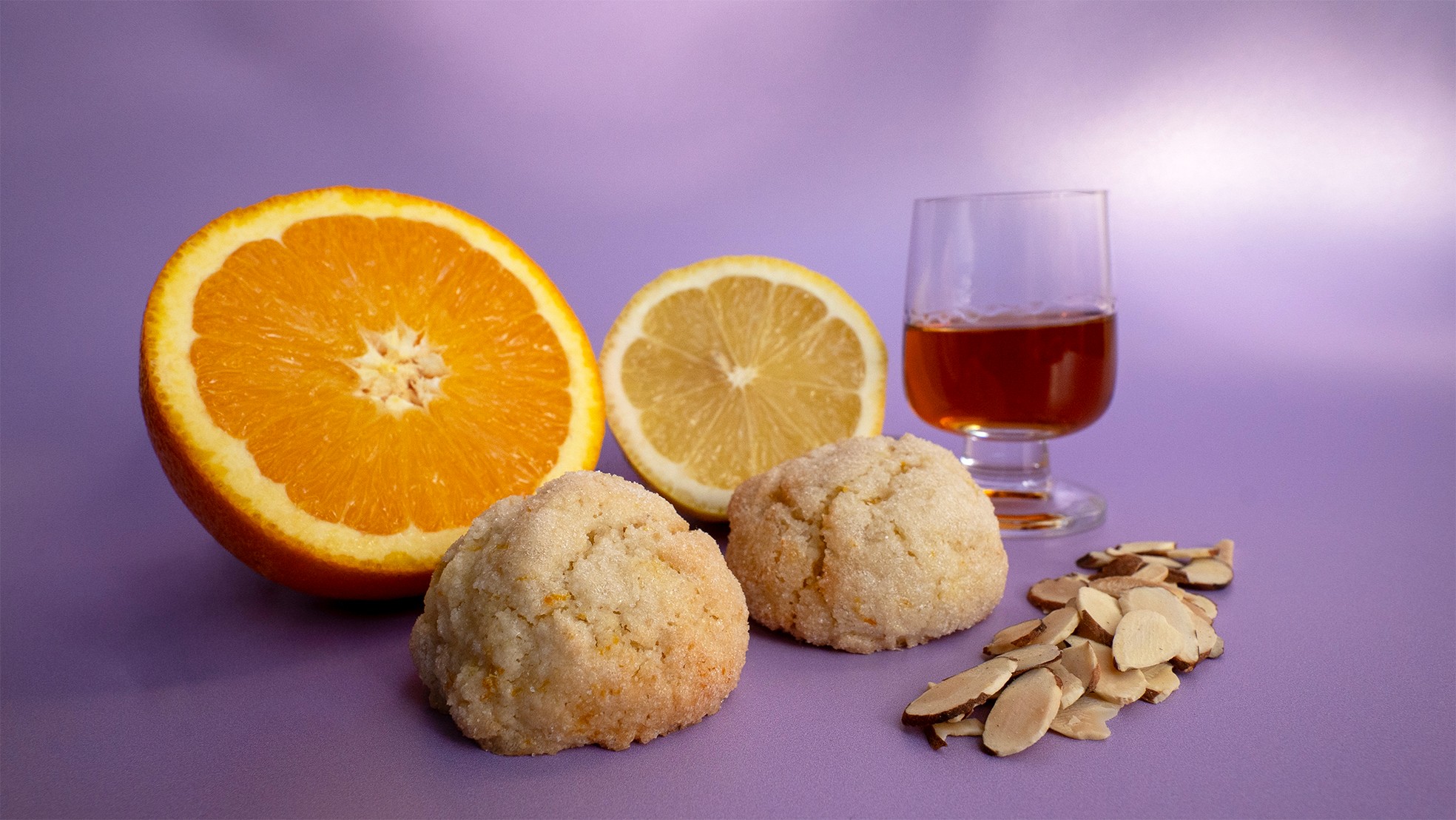 Half orange, lemon, two cauliflower florets, sliced almonds, and a glass of red liquid on purple background.