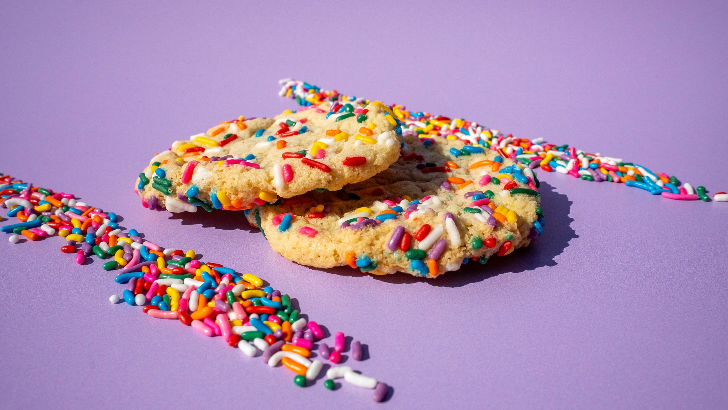 Three cookies with colorful sprinkles on a purple background, with sprinkles scattered around.
