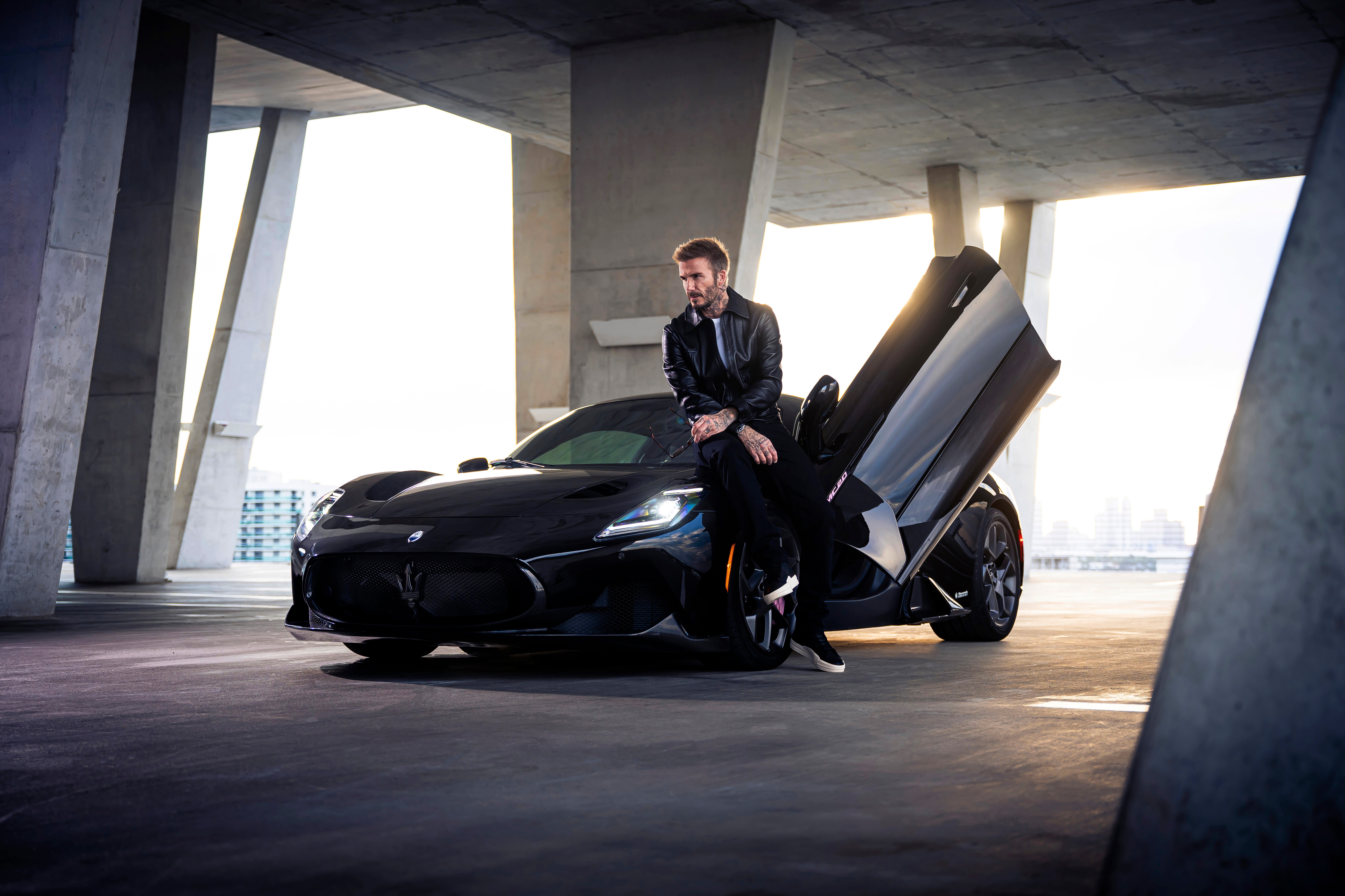 Man in a suit standing next to a black sports car with gull-wing doors open, in a modern concrete structure, city skyline in background.