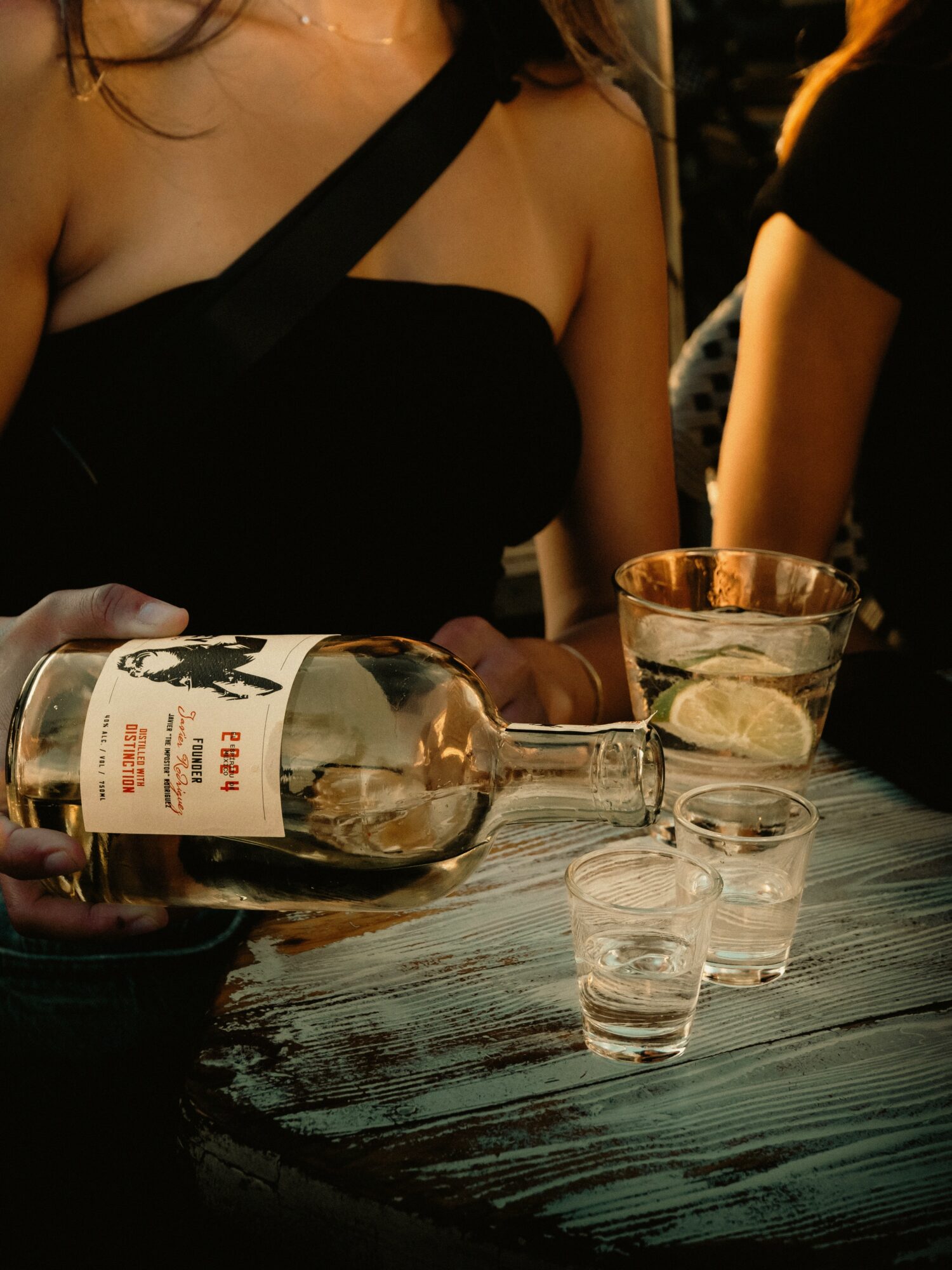 Person pouring clear liquid from a bottle into a glass on a dark wooden table, with another glass nearby.