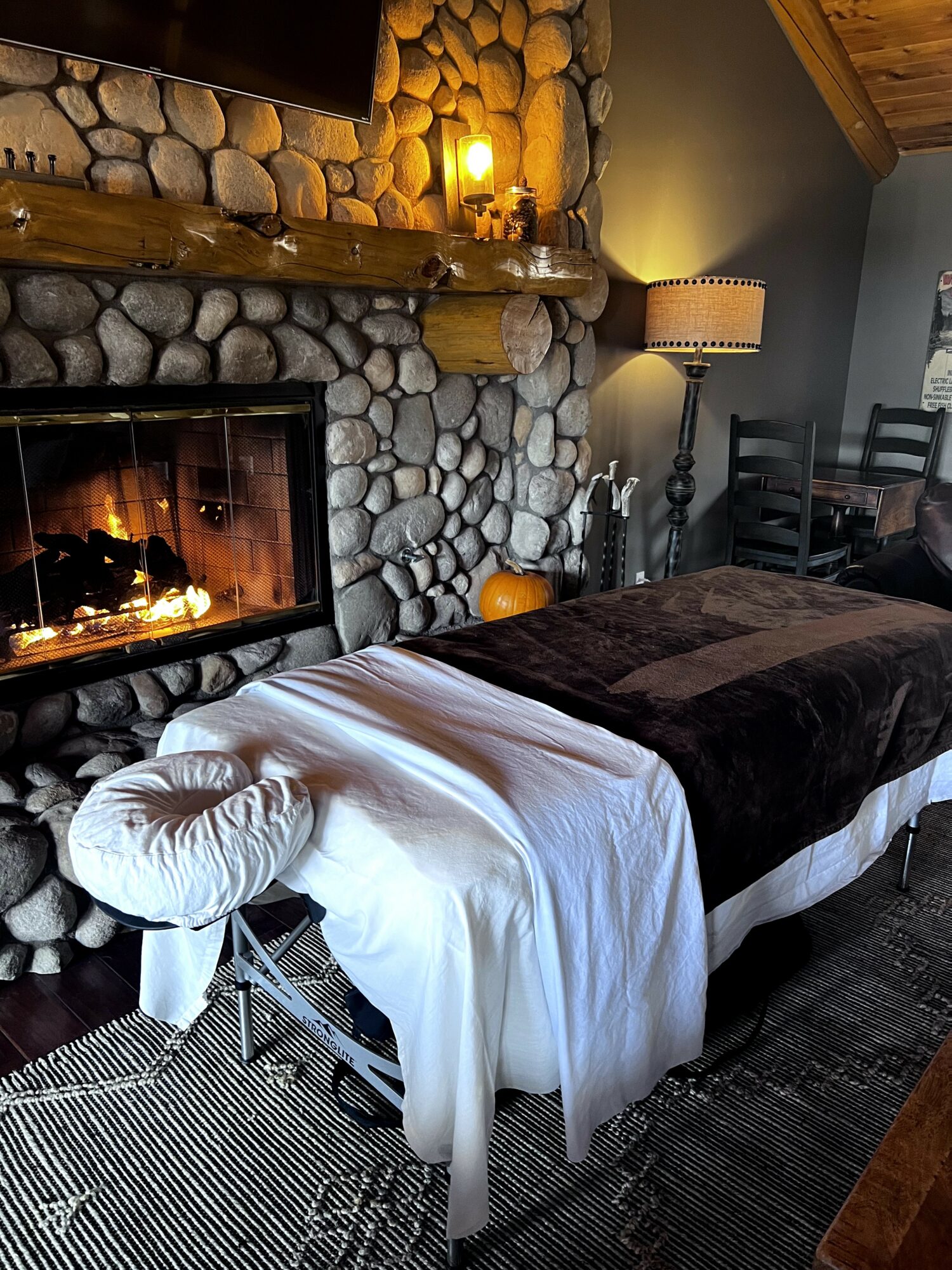 Massage table with white sheet and dark blanket in cozy room with stone fireplace, wooden mantel, and warm lighting.