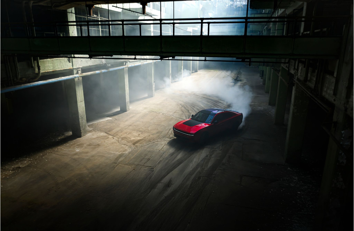 A red sports car drifting in an industrial setting with dust and smoke, under a bridge or overpass.