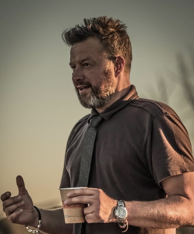 Man with beard and short hair holding a cup, wearing a dark shirt and watch, outdoors at sunset.