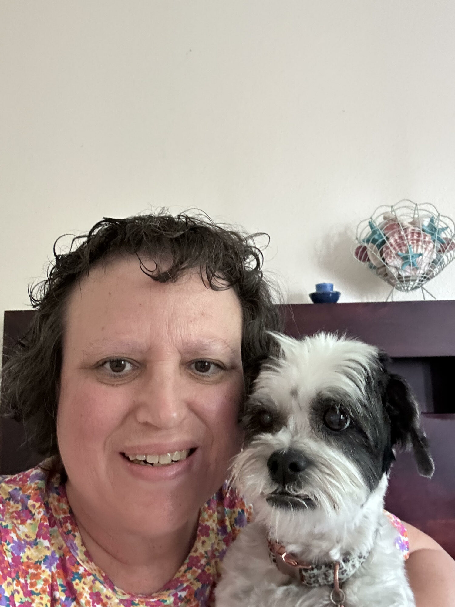 Person with short curly hair smiling next to a small white and black dog with a collar, indoors.