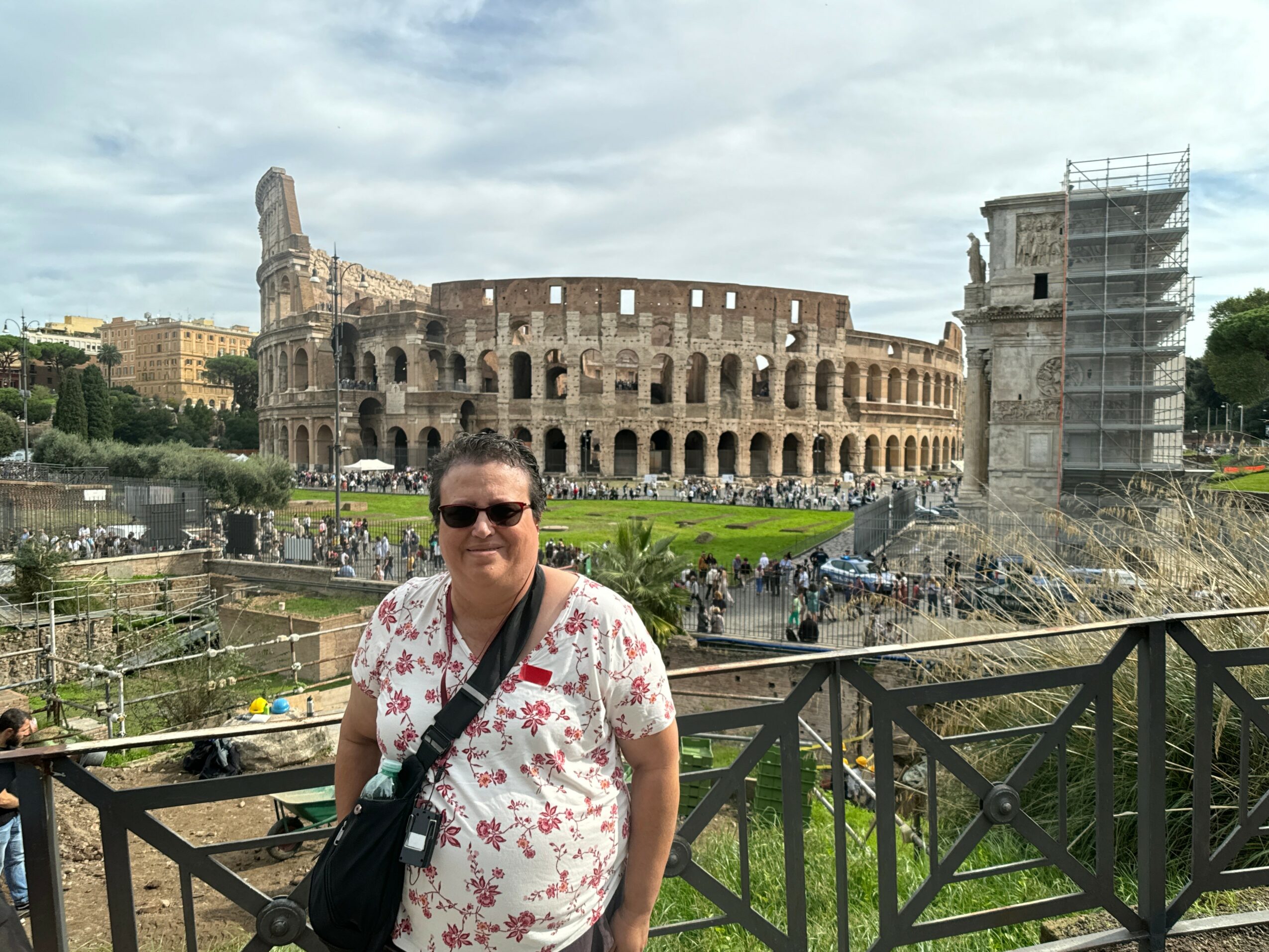 Person wearing sunglasses and a floral shirt standing in front of the Colosseum in Rome, Italy, with scaffolding on one side.