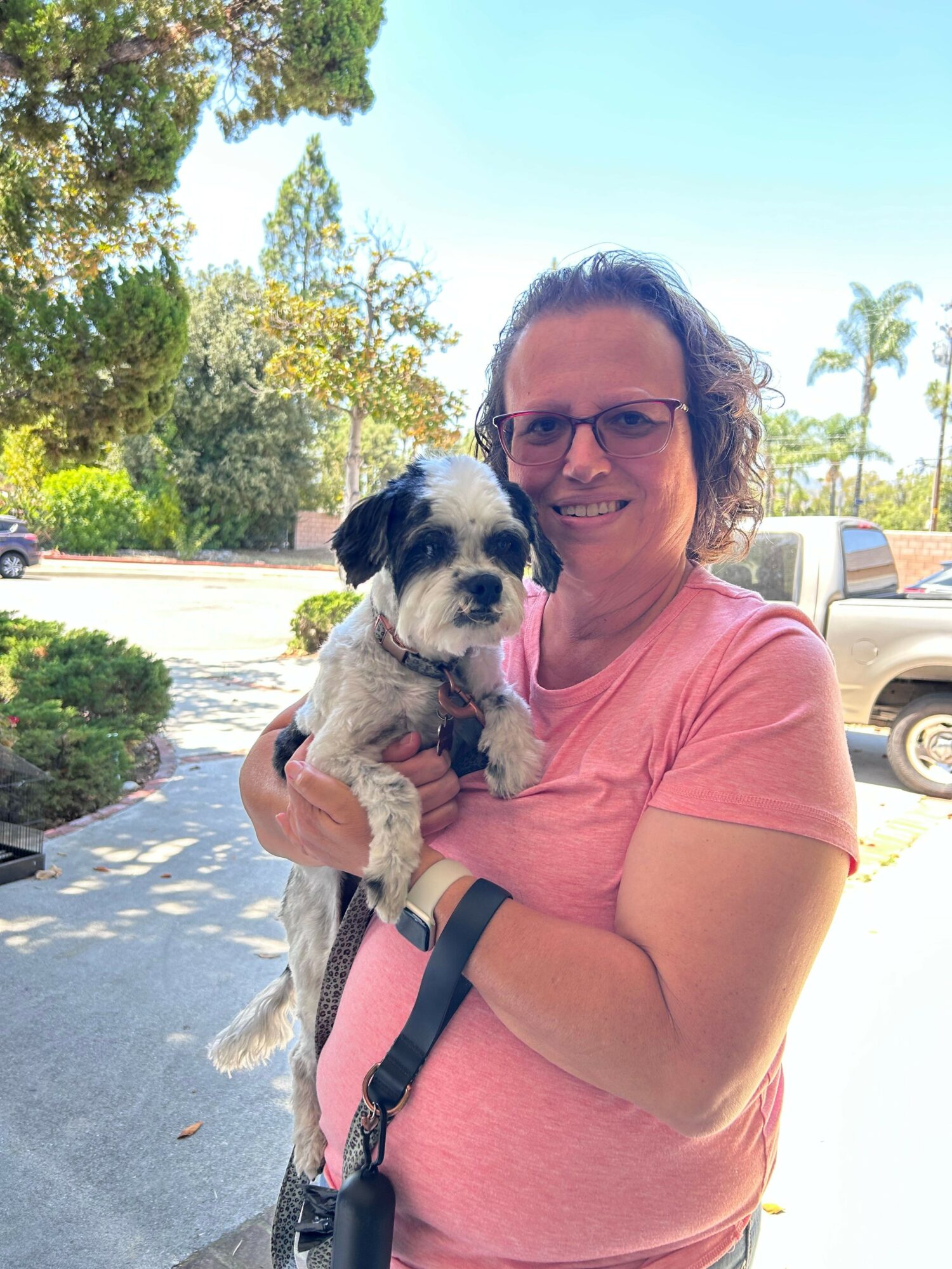 Woman holding a small dog outdoors, smiling, with trees and a car in the background.