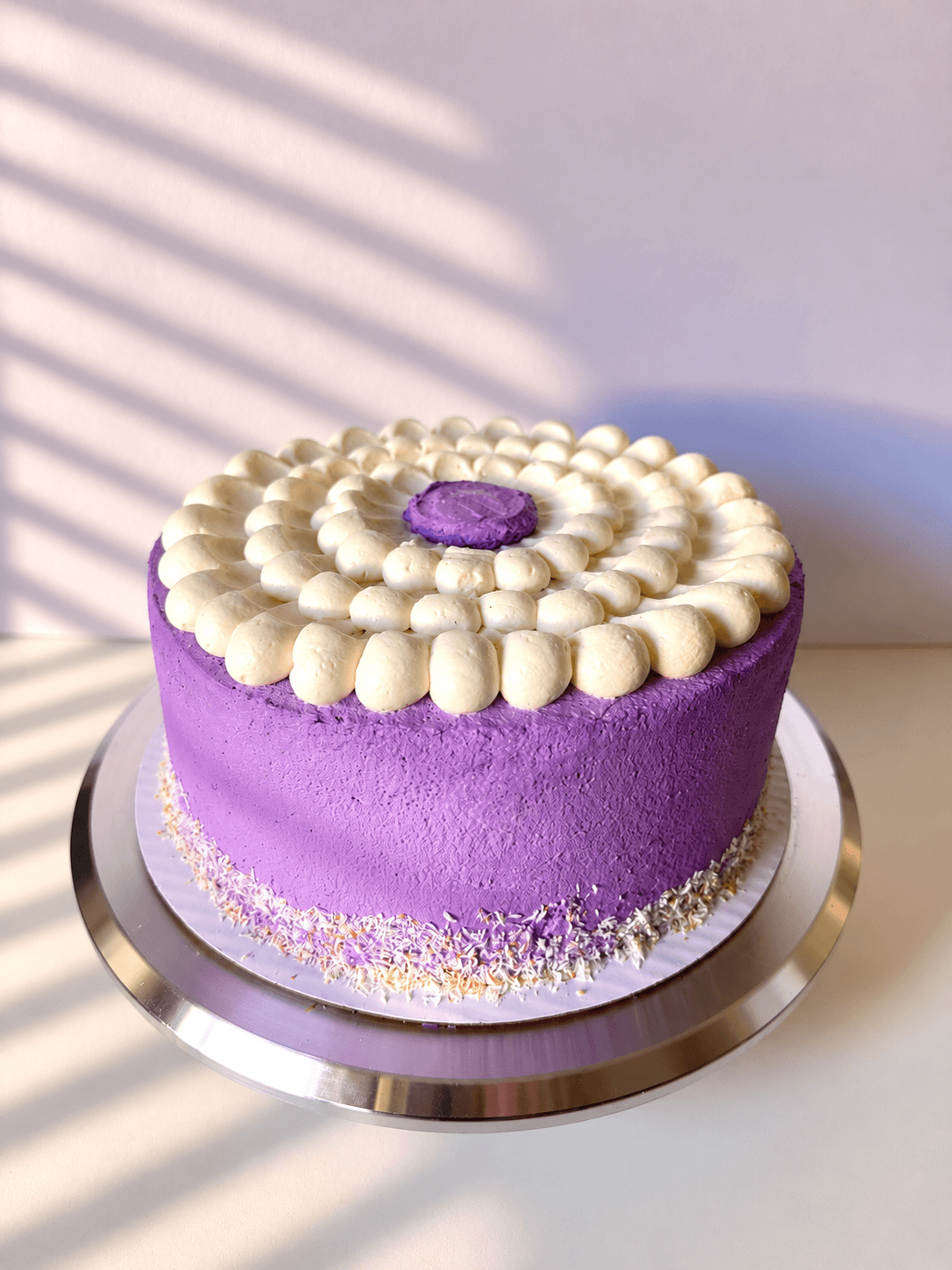 Round purple cake with white frosting decoration on top, on a silver cake board, with shadows cast on a light background.