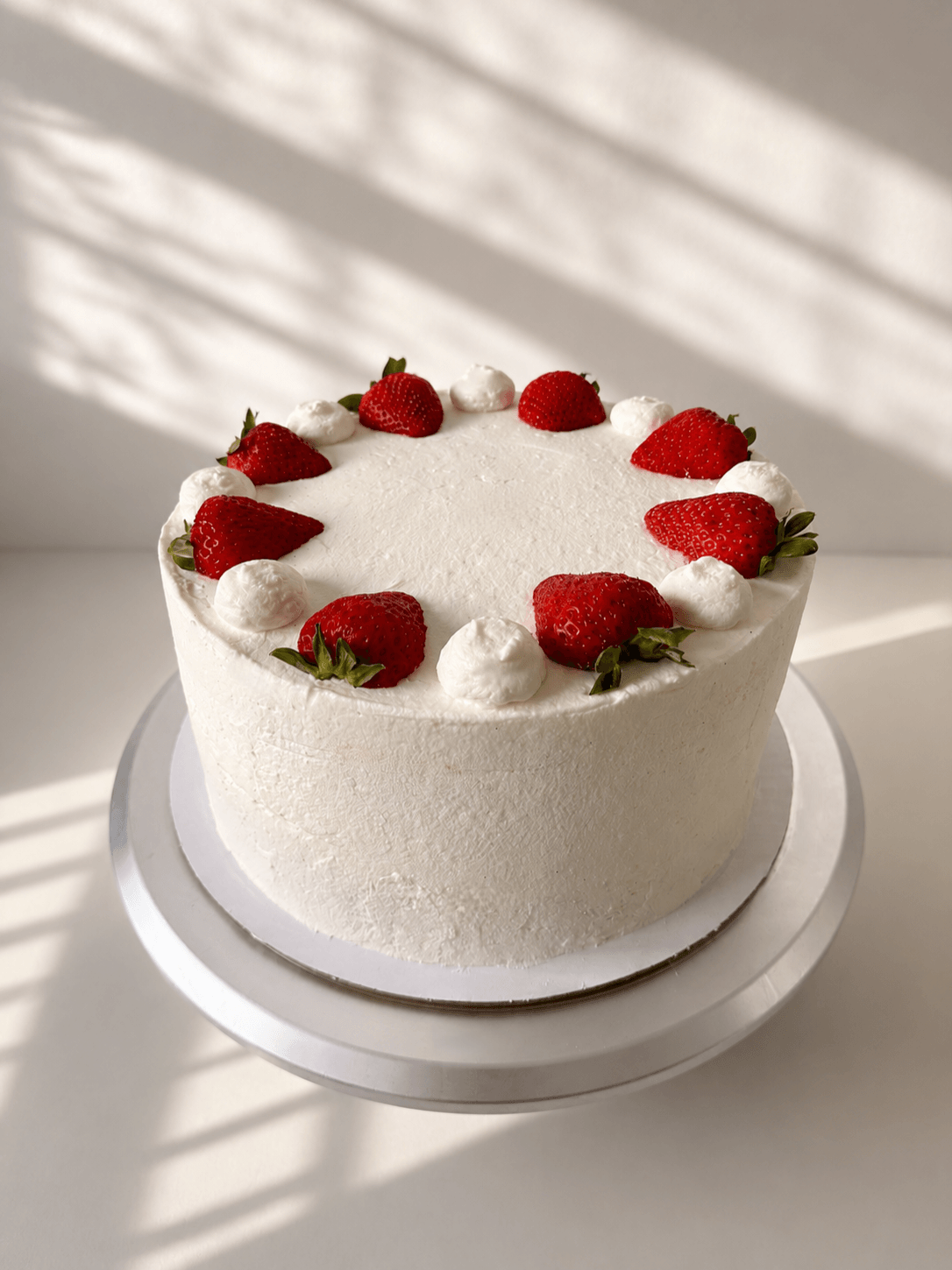 Round white cake with strawberries and dollops of cream on top, on a white surface with shadows, minimal background.