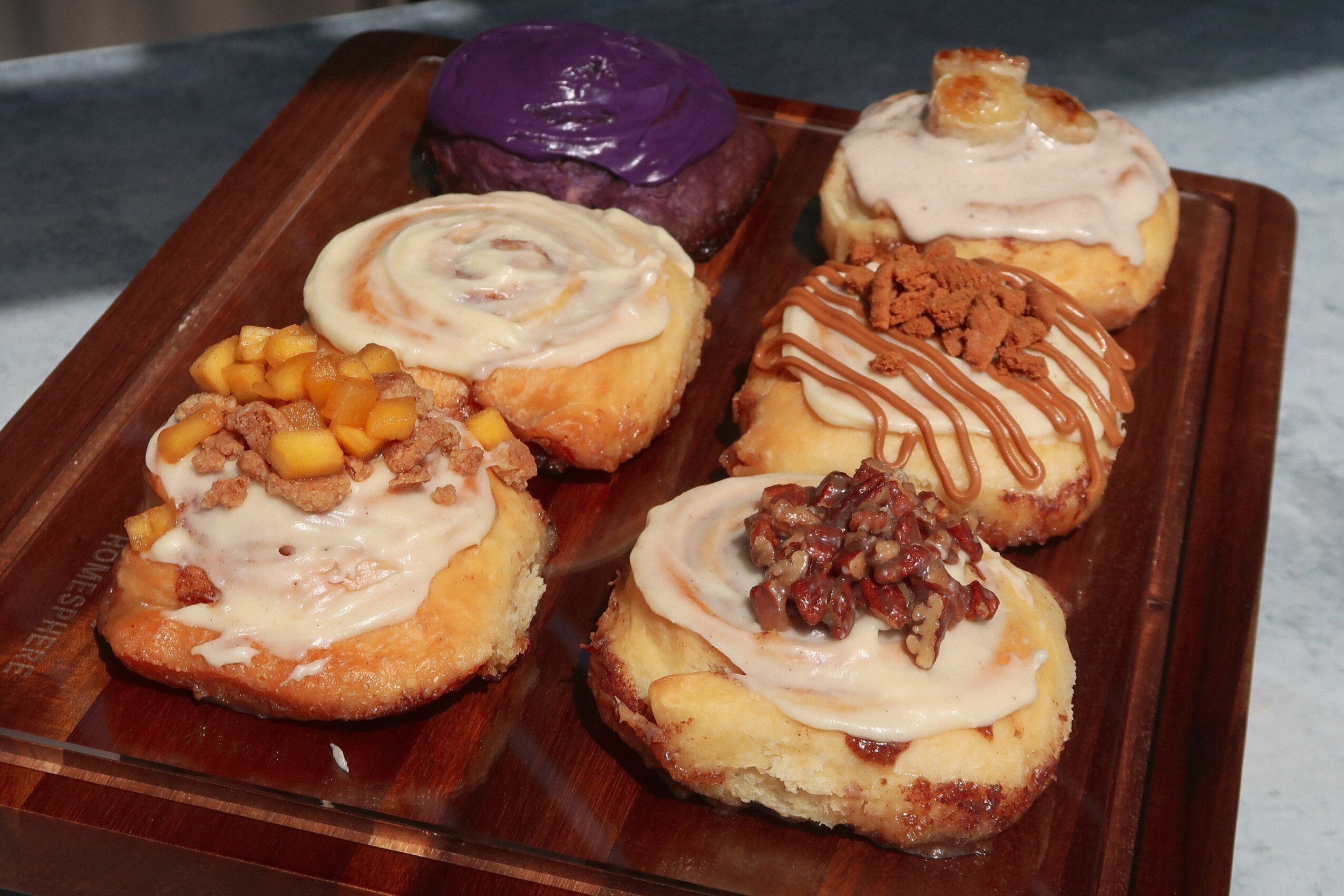 Six assorted mini donuts with various toppings on a wooden tray, including purple glaze, caramel, chocolate drizzle, and fruit bits.