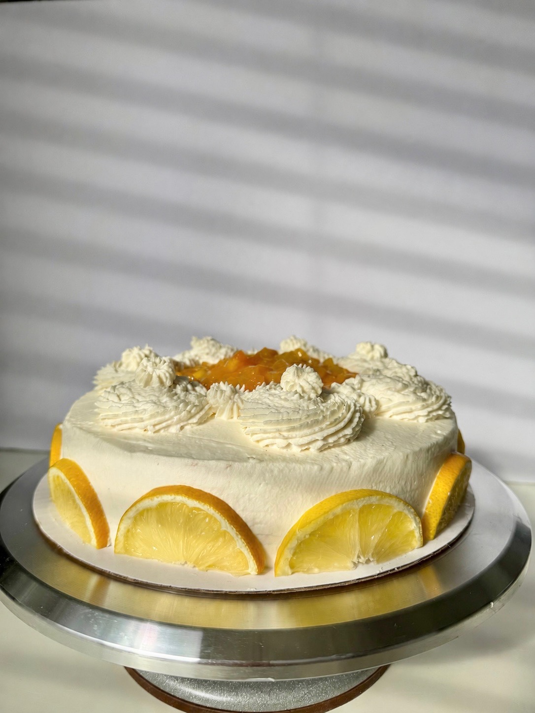 Round cake with white frosting, lemon slices around the edge, and a fruit topping in the center, on a silver stand.