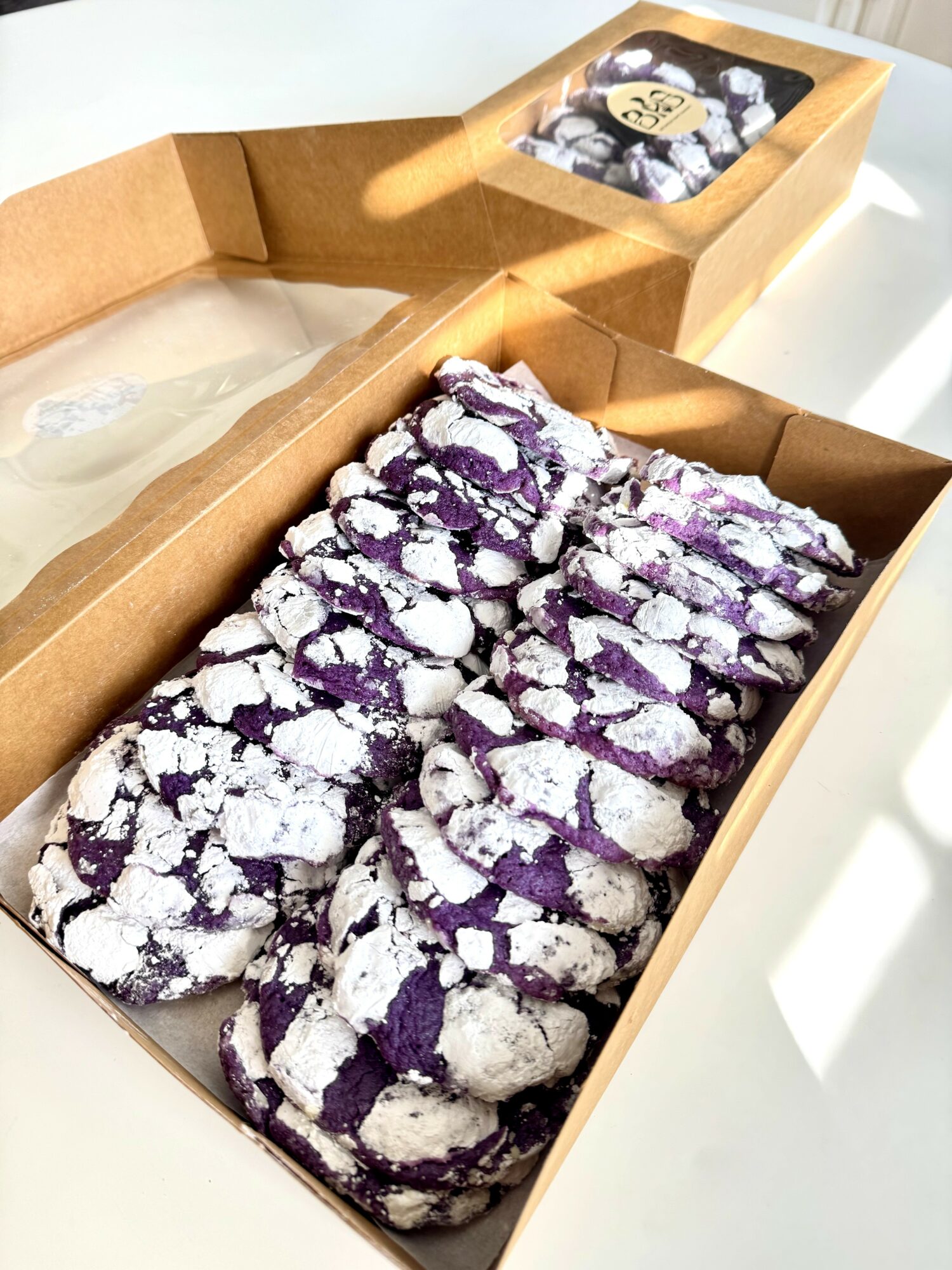 Purple and white cookies dusted with powdered sugar in a cardboard box, with a smaller box of cookies in the background.