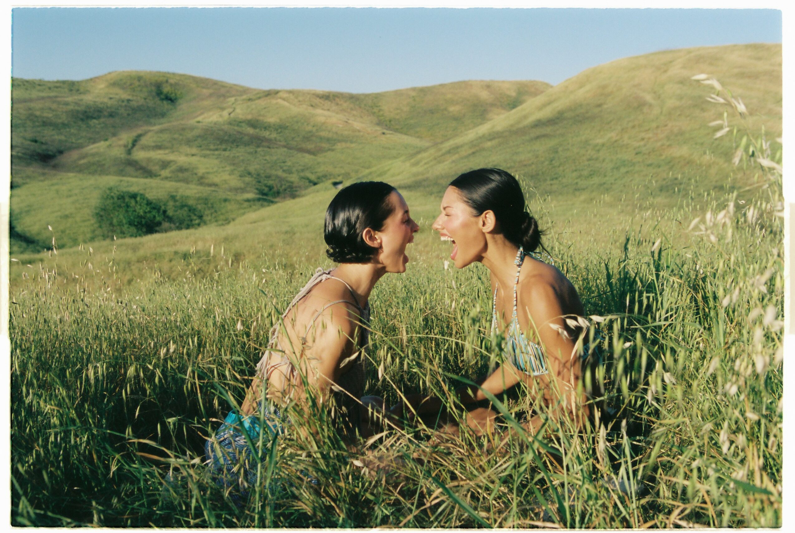 Two women face each other in a grassy field with rolling hills in the background, smiling and leaning toward each other.