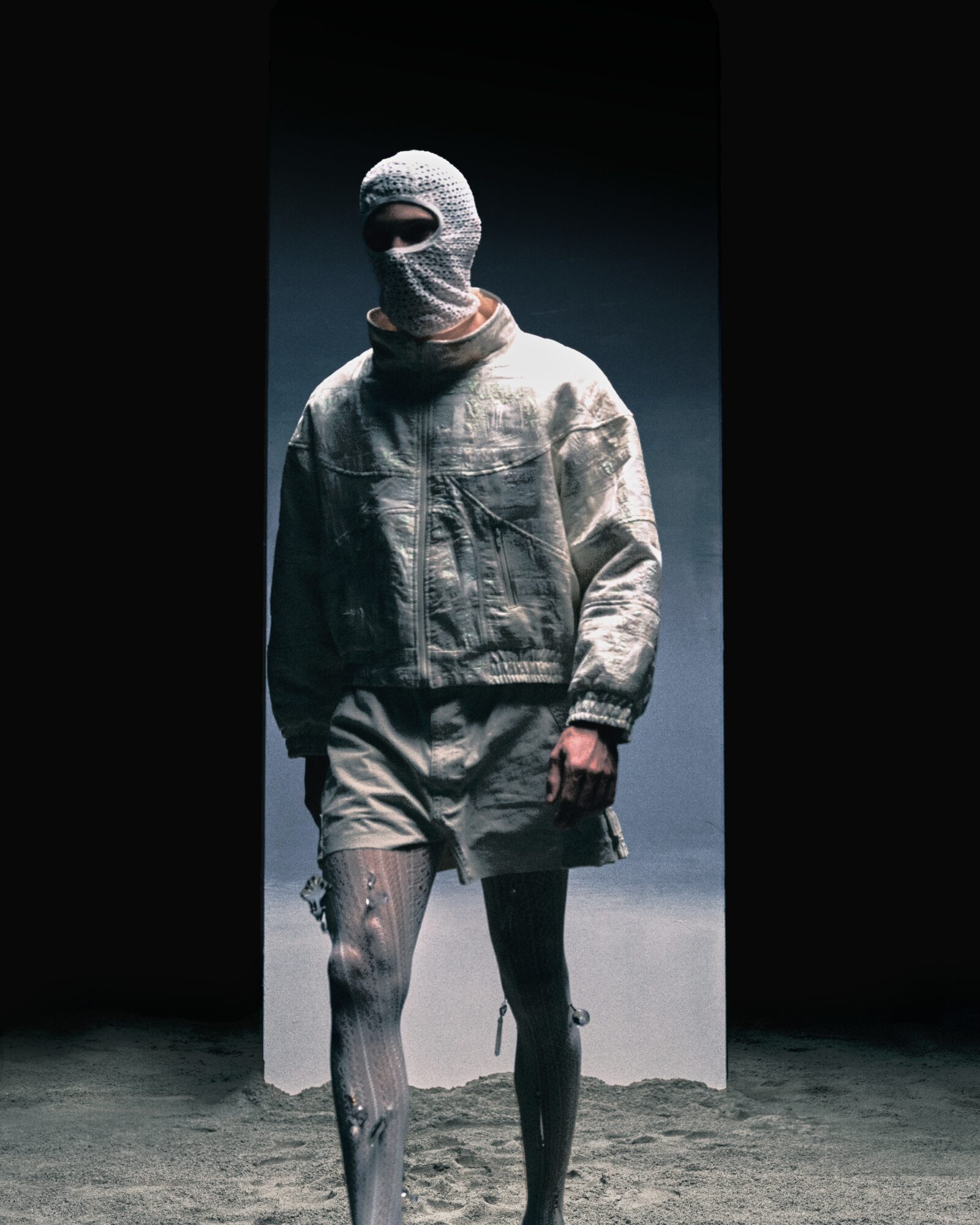 Person wearing a hooded mask and casual clothing, standing on a runway with a dark background.