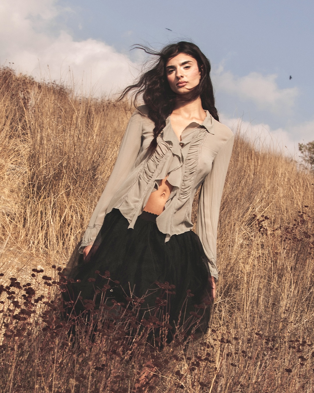 Young woman standing in a grassy field with a blue sky background, wearing a beige blouse and black skirt.