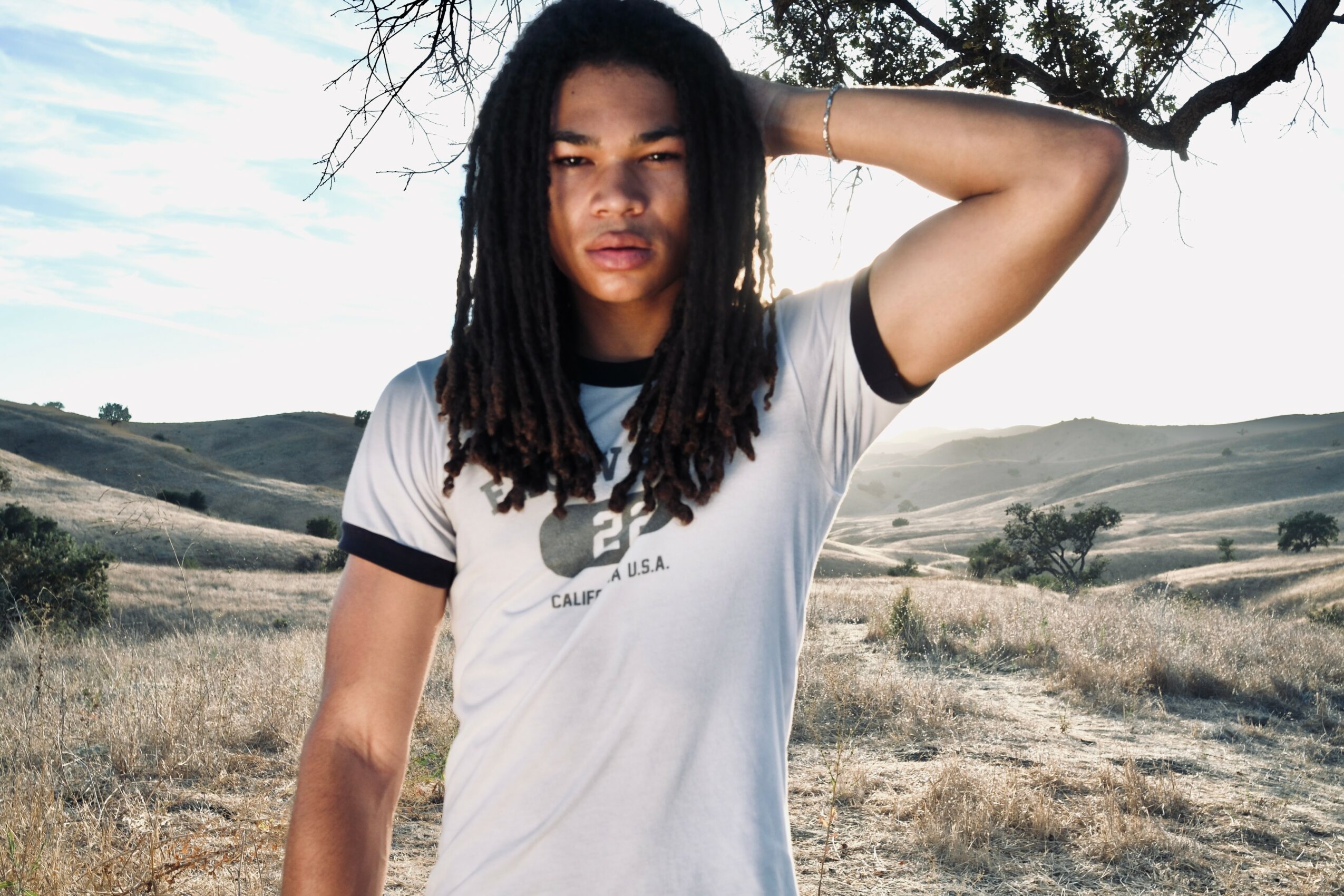 Young woman with dreadlocks standing outdoors in a landscape with rolling hills and trees, wearing a white T-shirt with black trim, arm raised behind her head.
