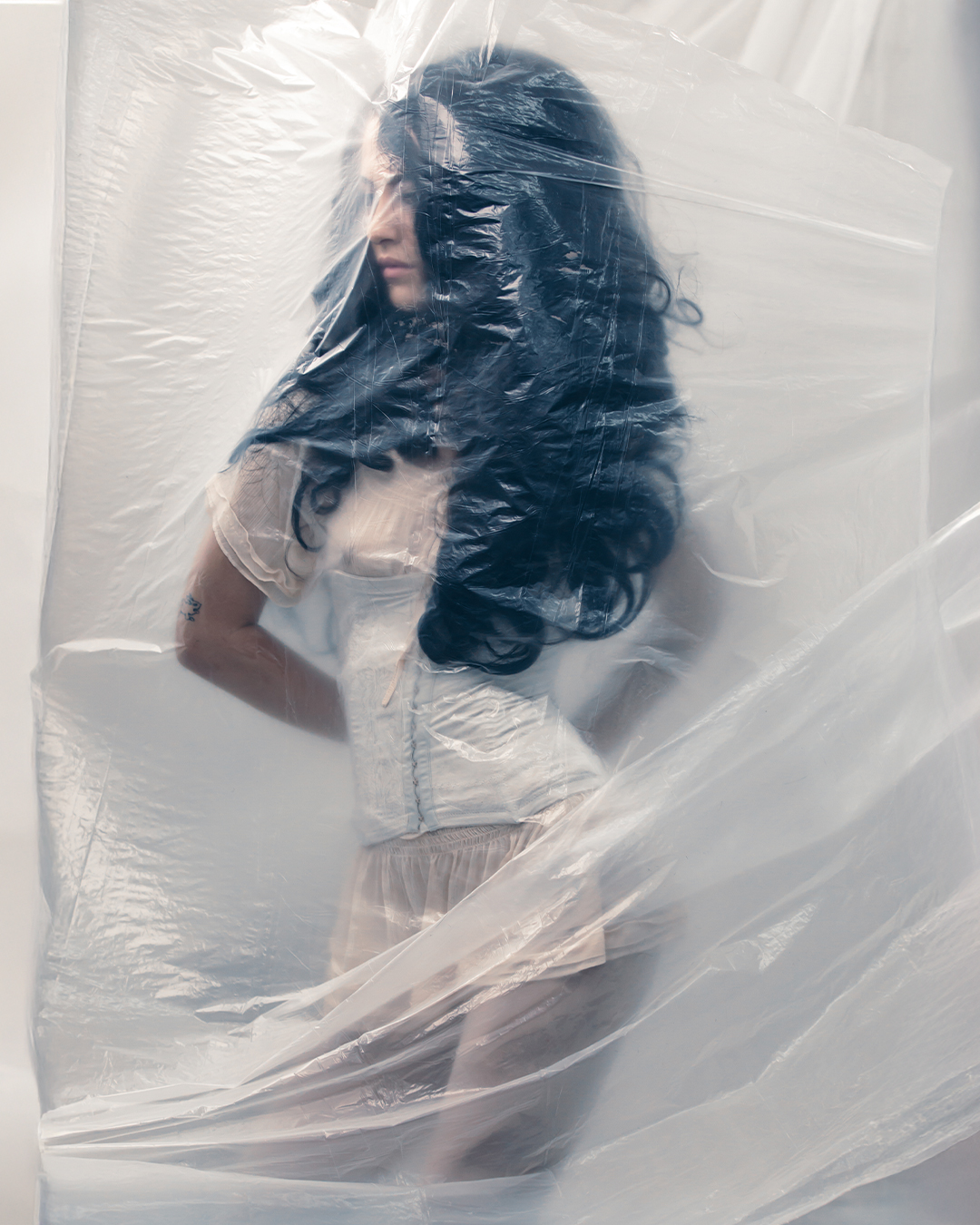 Woman with long, wavy hair standing behind translucent plastic, facing left, with her eyes closed and arms crossed.