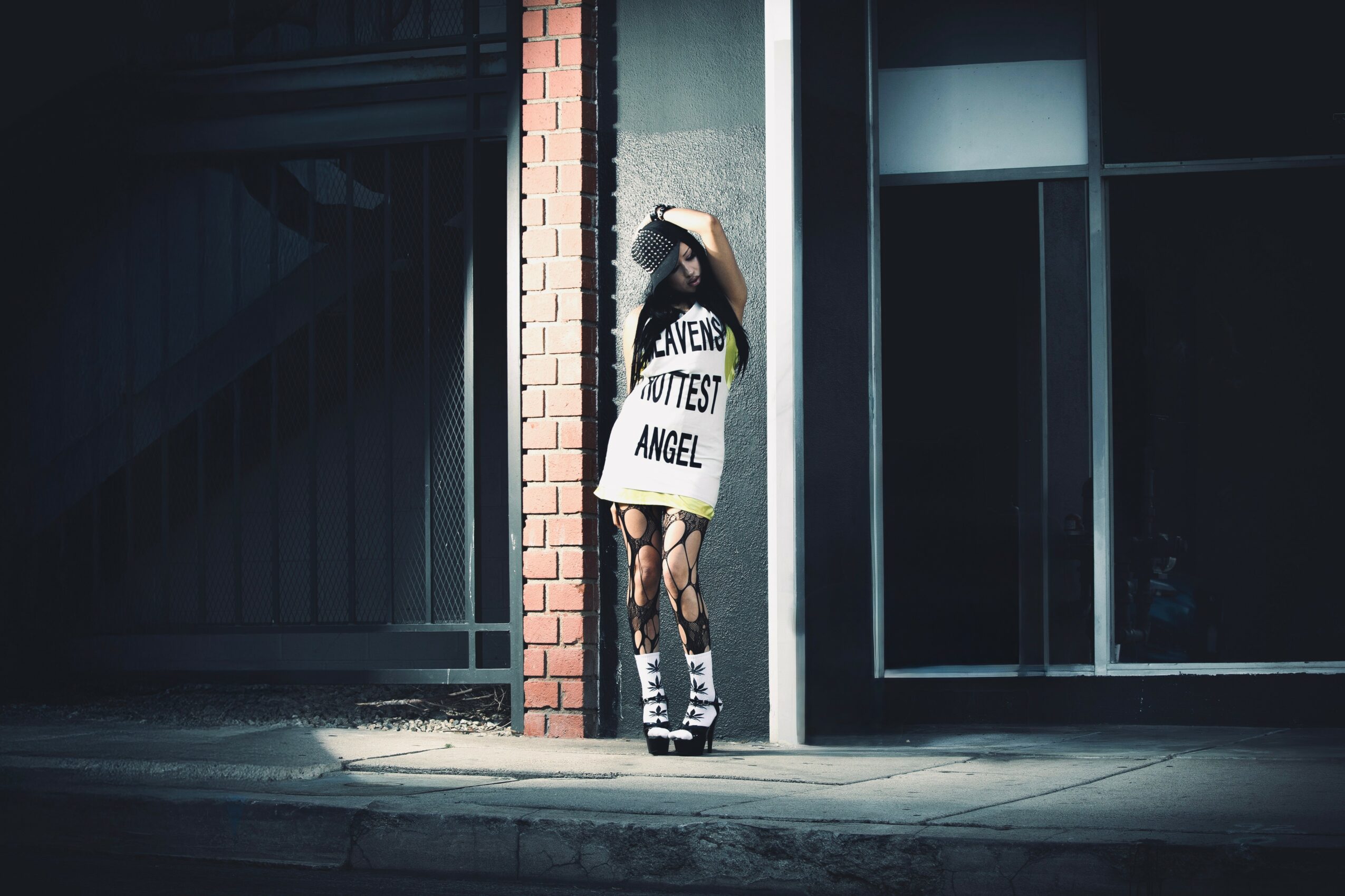 Person standing against a wall with a sign reading 'WITCHIEST ANGEL', wearing a hat and striped socks, in an urban setting.