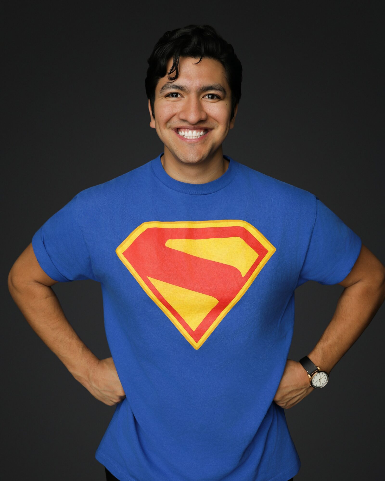 Person smiling, wearing a blue Superman T-shirt with a red and yellow emblem, standing with hands on hips against a dark background.