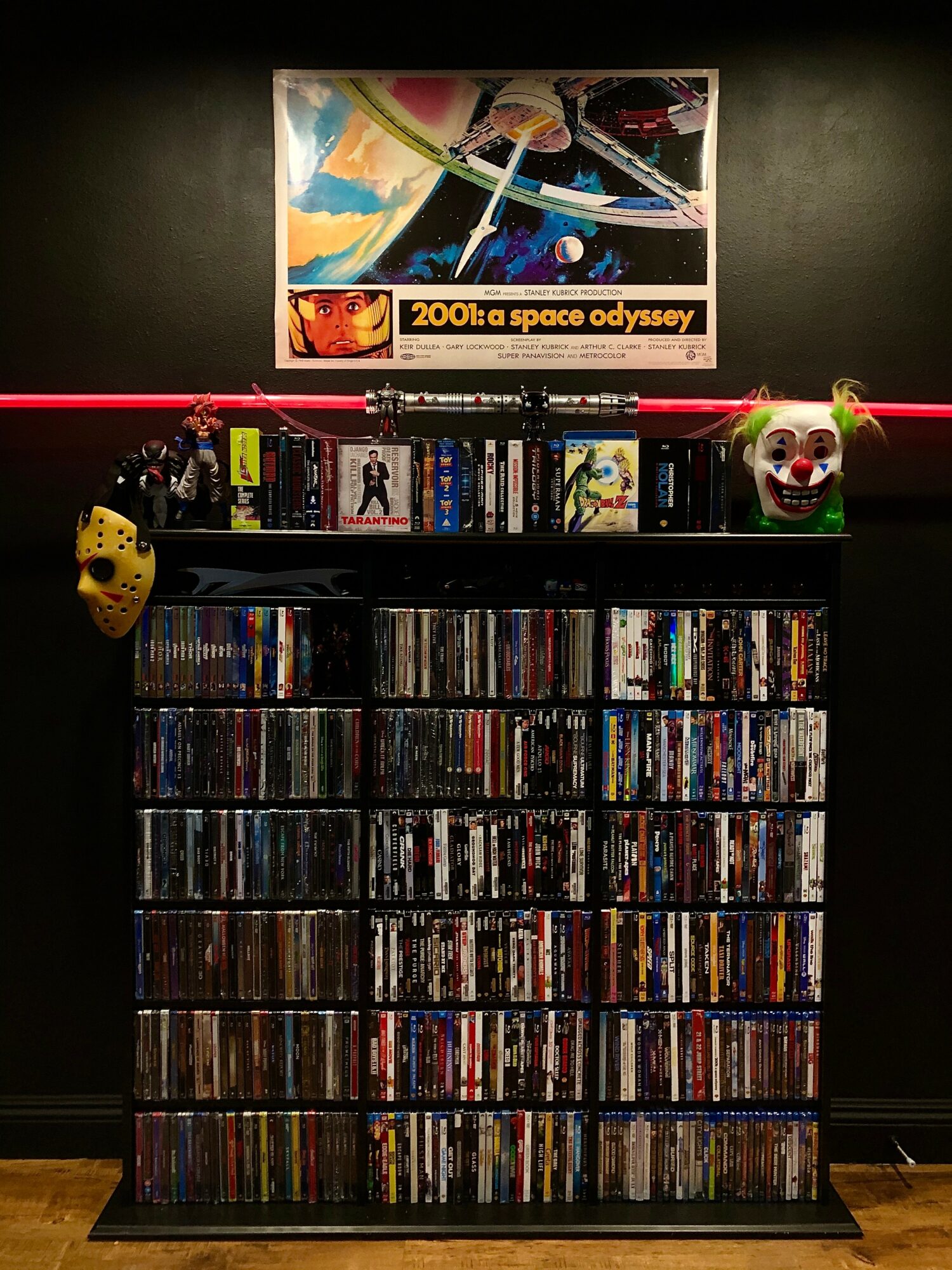 Bookshelf with DVDs, a clown mask, and a yellow object, against a black wall with a space-themed poster above.