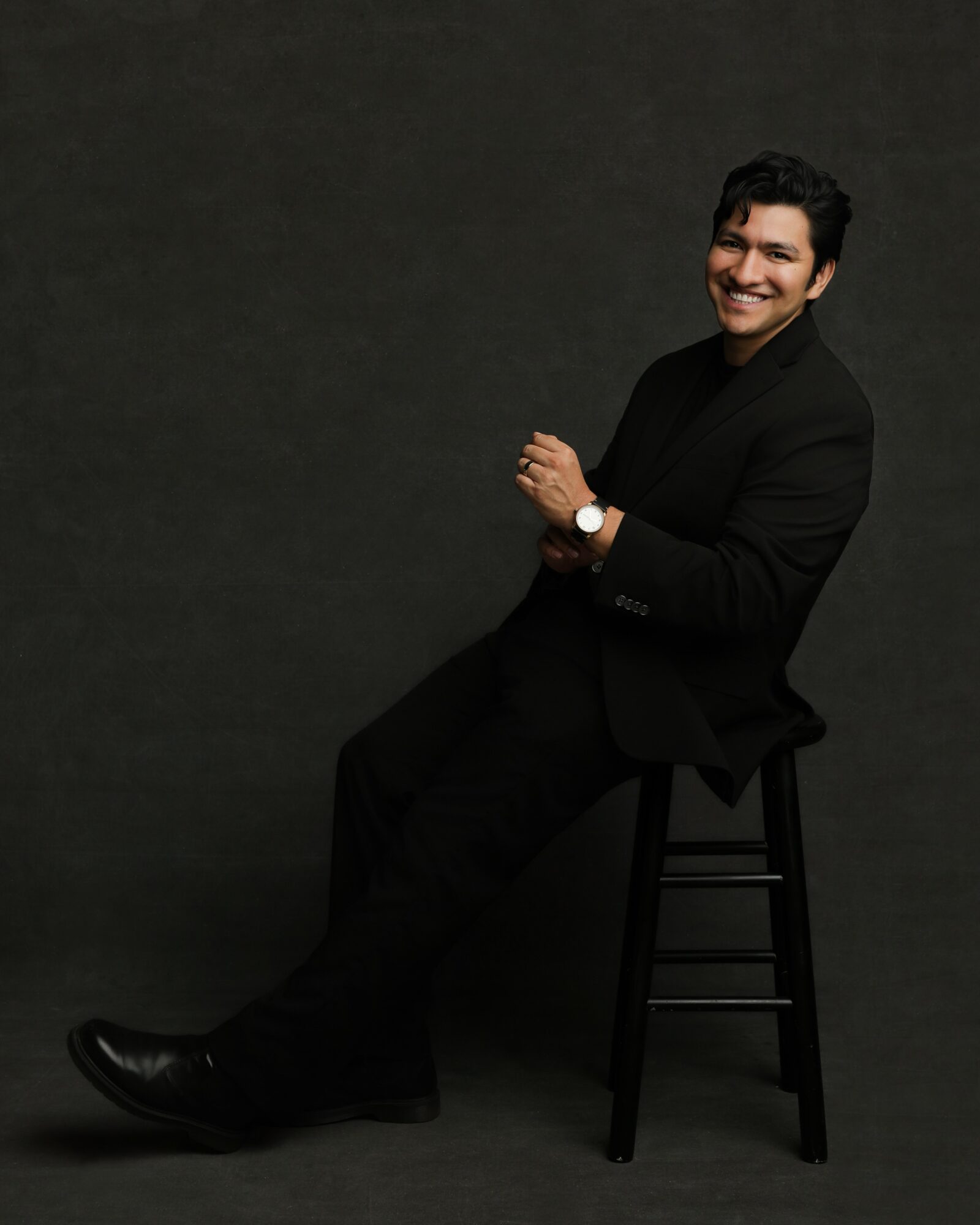 Man in black suit sitting on a stool, smiling, with a dark background.