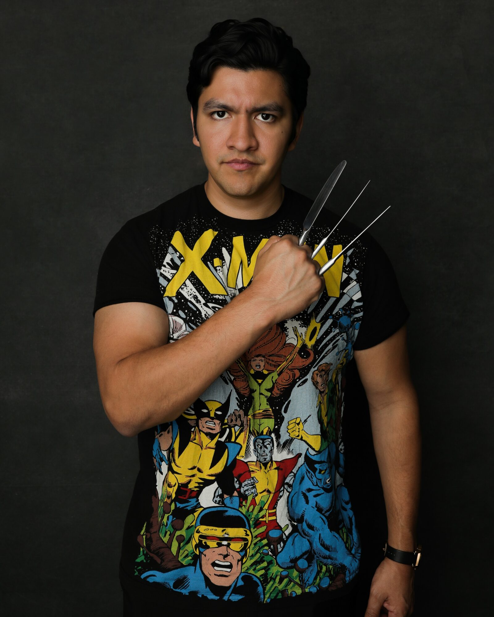 Young man with black hair holding three metal claws, wearing a colorful comic book-themed T-shirt, against a dark background.