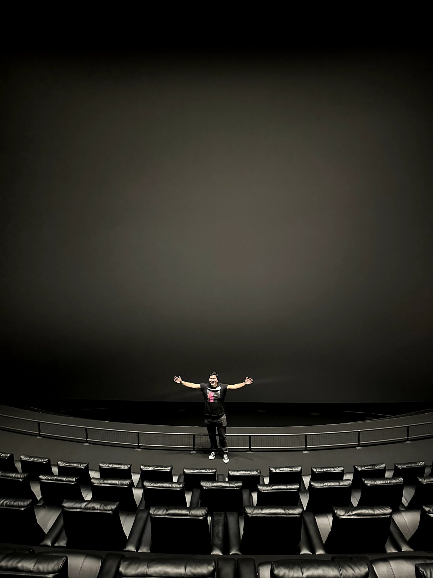 Person standing on stage with arms outstretched in an empty theater, large dark screen behind, empty seats in front.