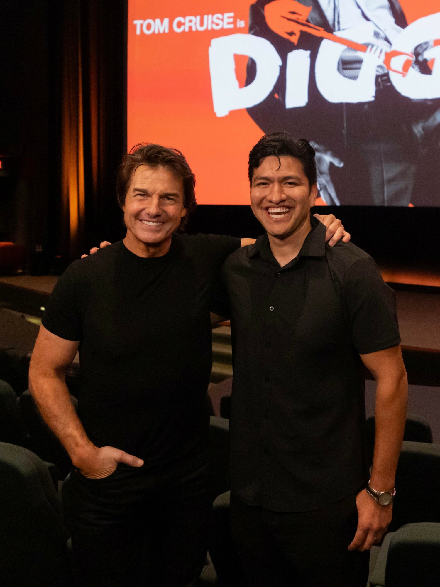 Two men smiling and standing together in a theater, with a movie poster in the background.