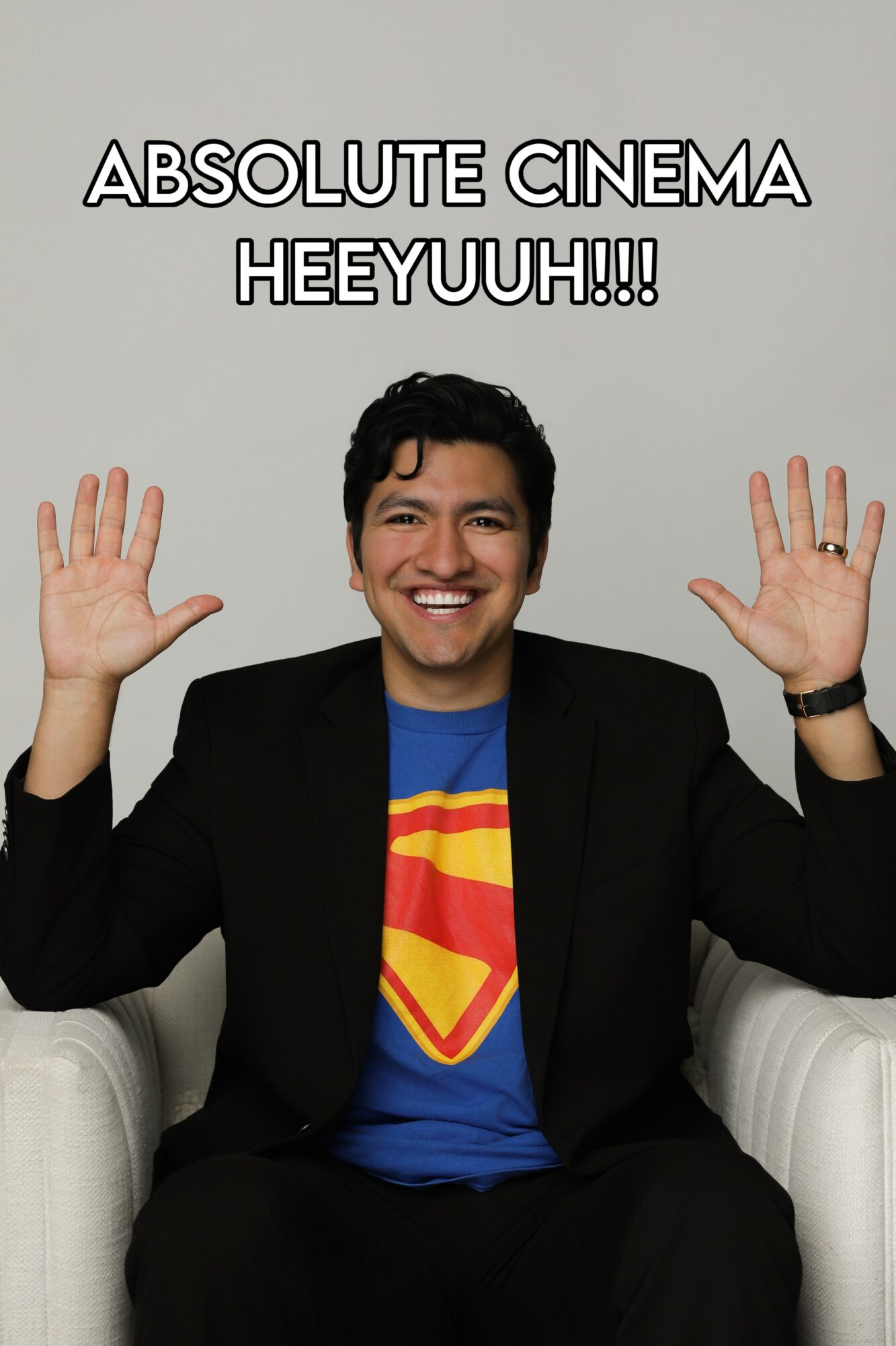 Man smiling with hands raised, wearing a Superman shirt and black blazer, sitting on a white chair, text above.