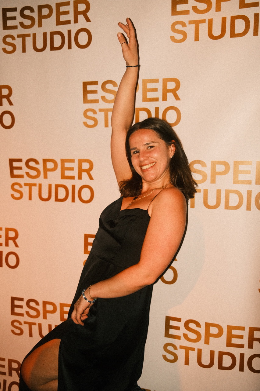Smiling woman in black dress with one arm raised, standing in front of a backdrop with 'ESPER STUDIO' text.