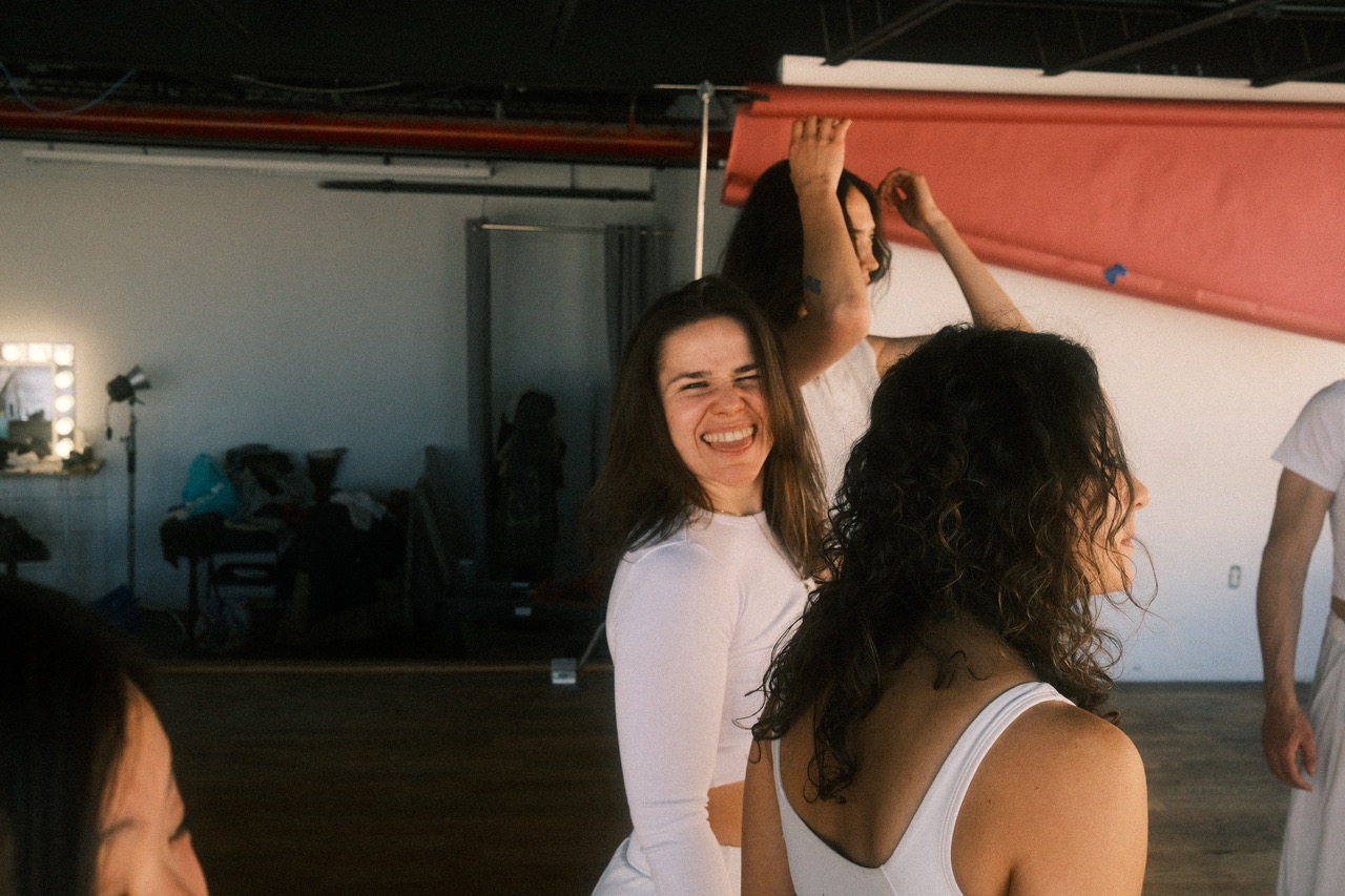 Two women smiling and laughing, one with long dark hair and the other with curly hair, in a studio setting.