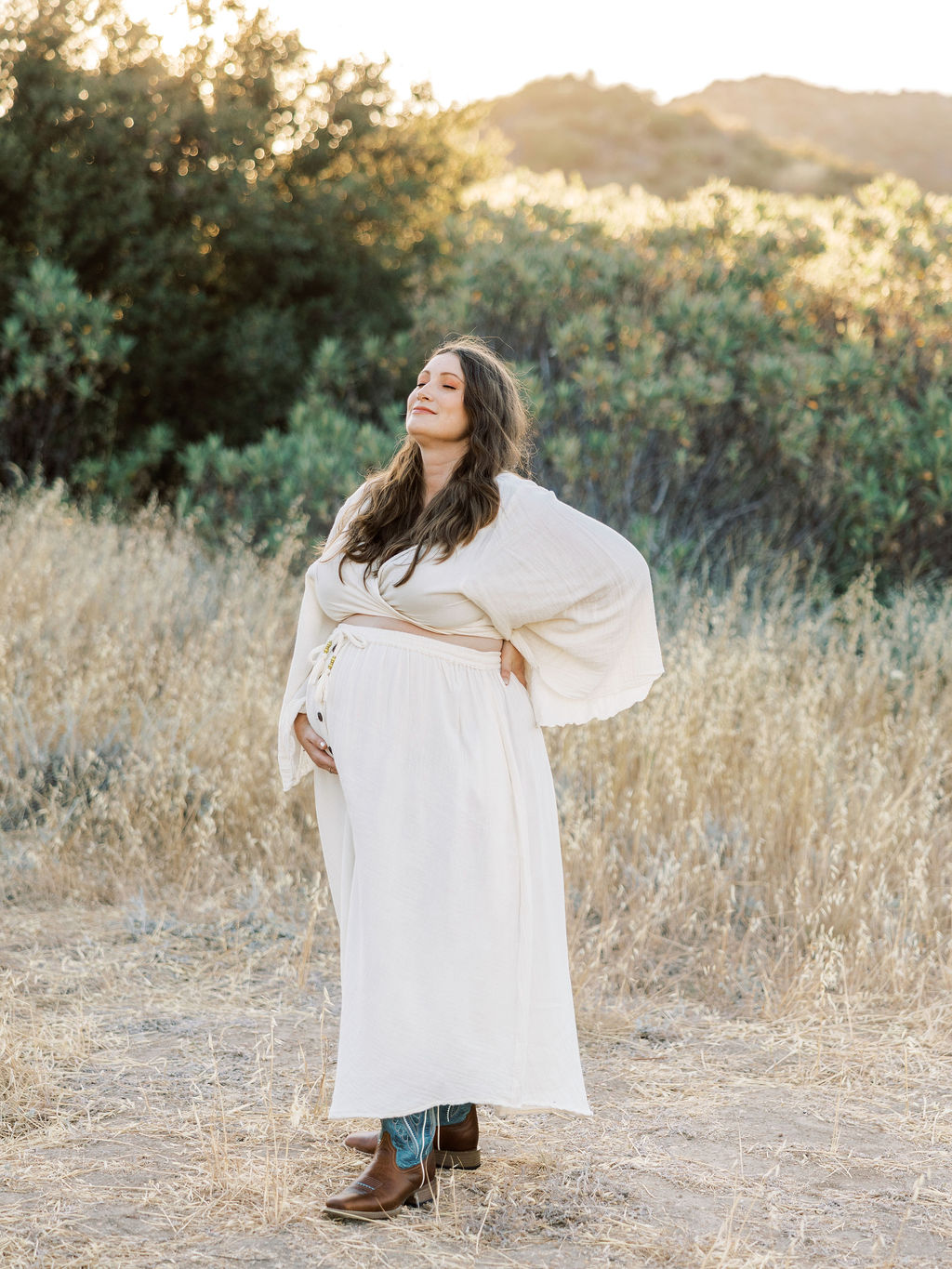 Woman standing outdoors in a field with trees, wearing a white dress and boots, looking up with closed eyes.