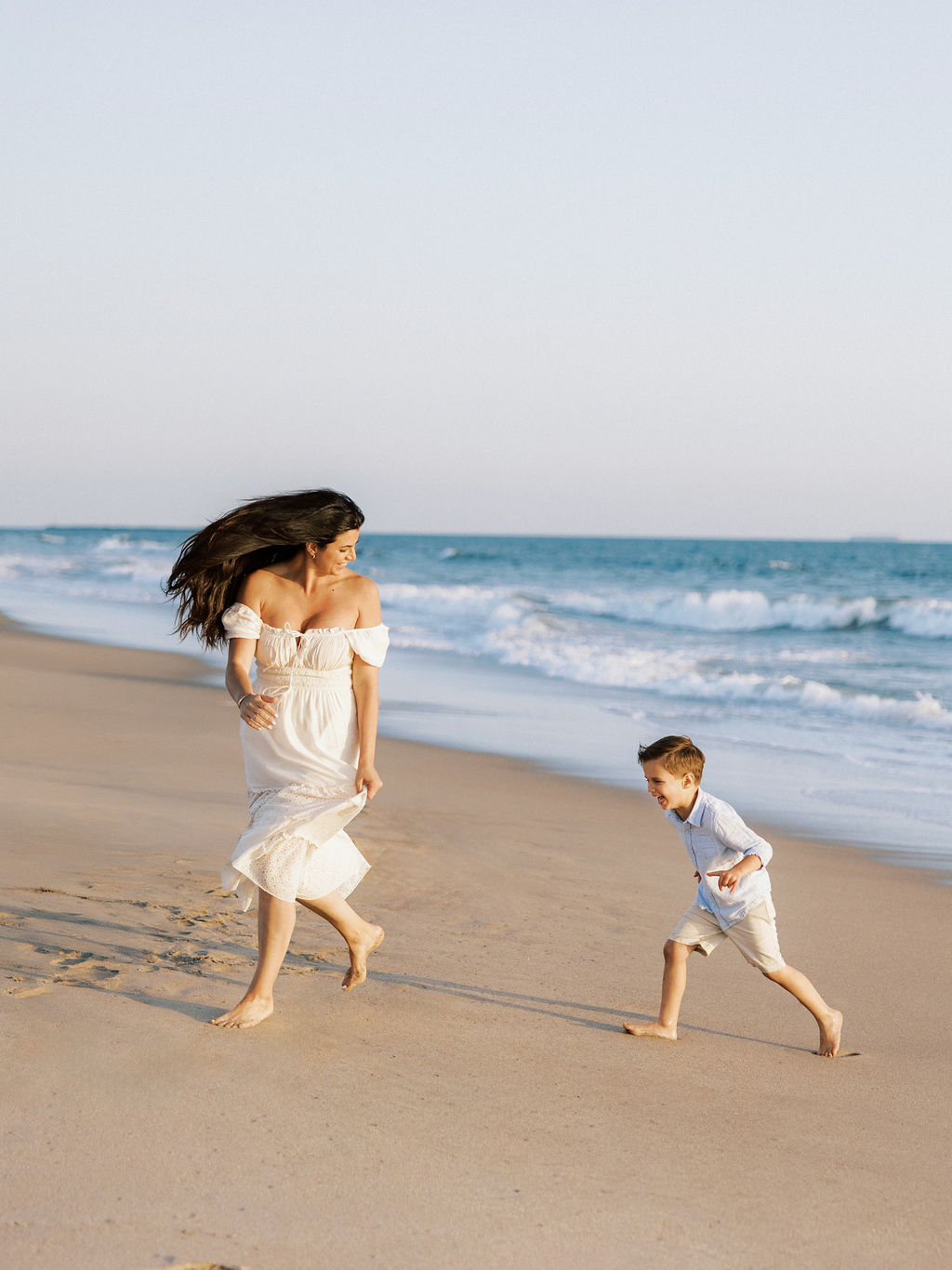 Woman and child walking and running on a sandy beach near the ocean, with waves in the background.