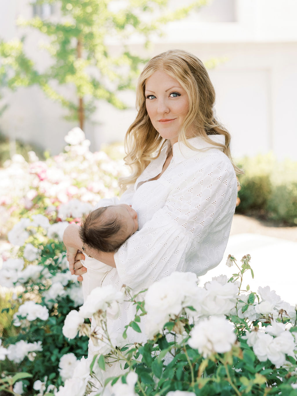 Woman holding a baby in a garden with white flowers, sunlight, and greenery, smiling gently at the camera.