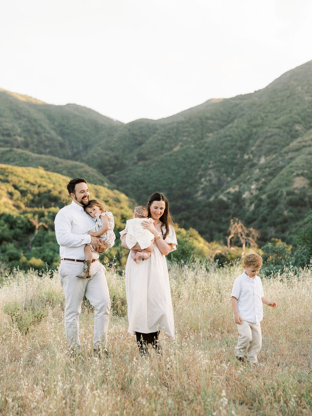 Family of four standing in a grassy field with mountains in the background, two adults and two children, during daytime.
