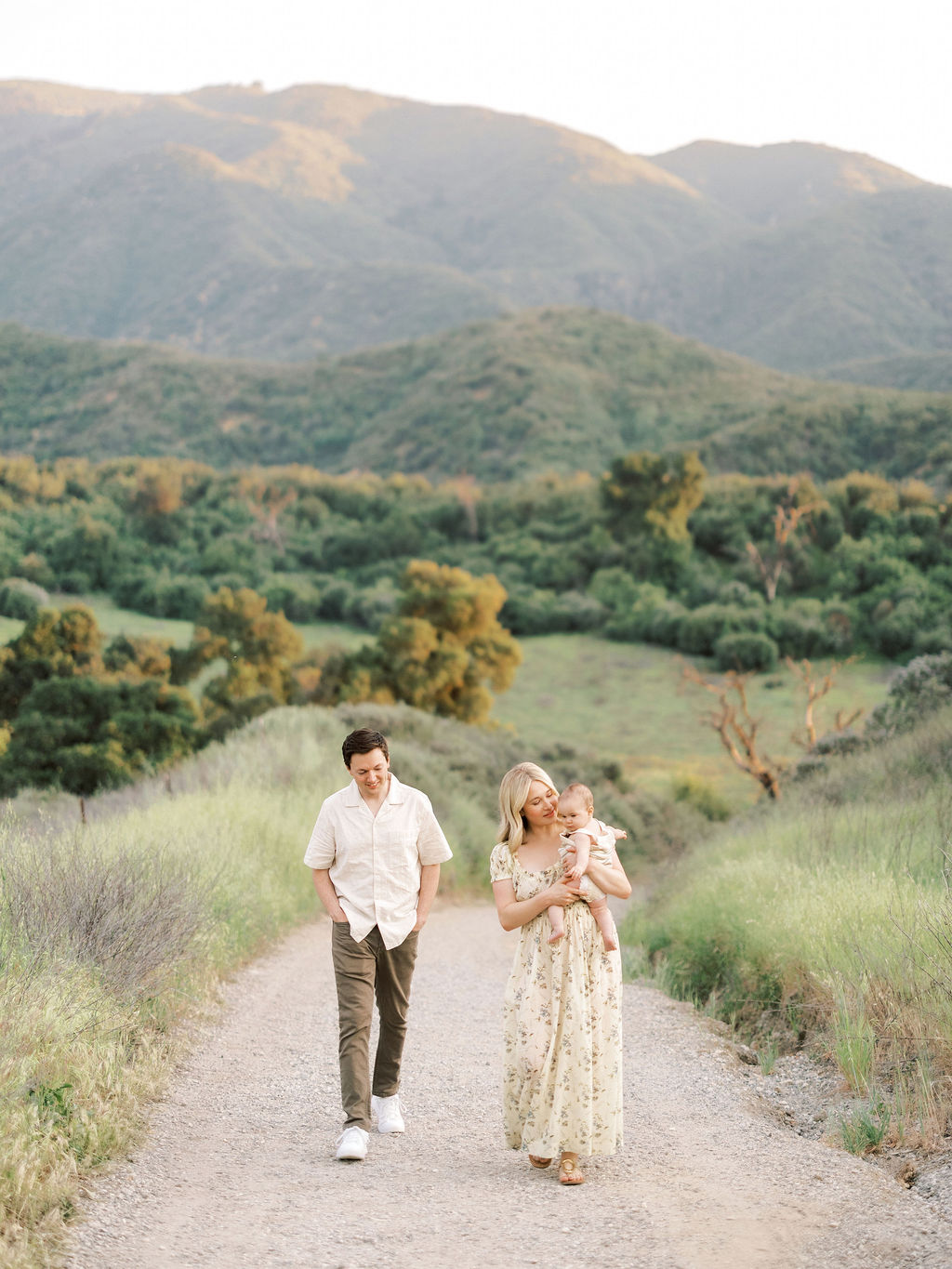 A couple walking on a dirt path in a green landscape with mountains in the background, woman holding a small dog.