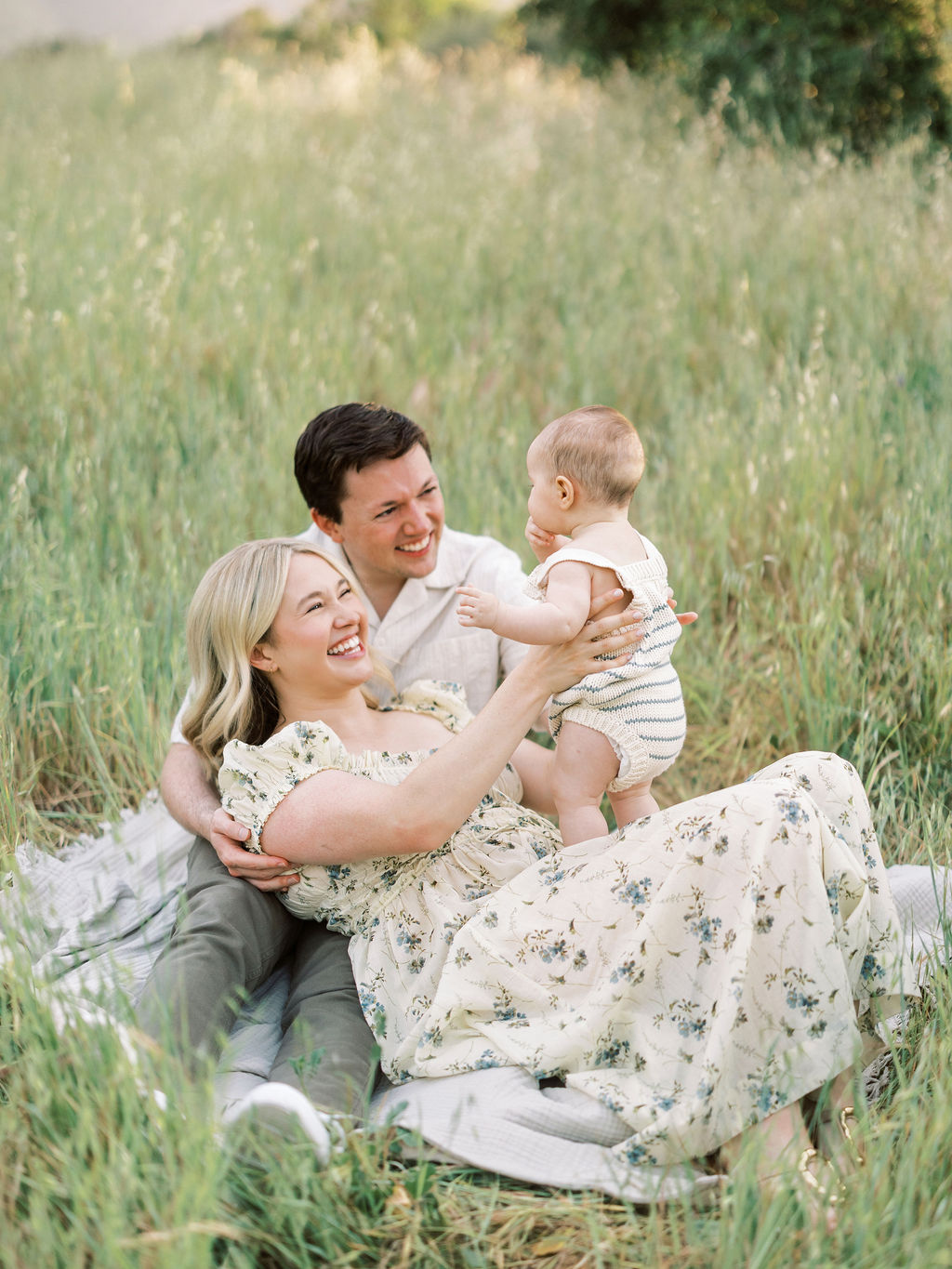 Family of three sitting in a grassy field, smiling and playing together outdoors.