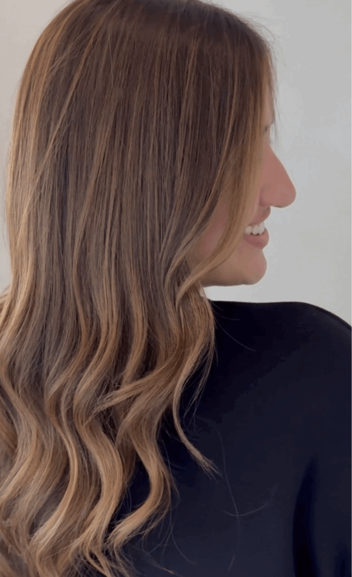 Side profile of a woman with long, wavy brown hair smiling, wearing a black top.