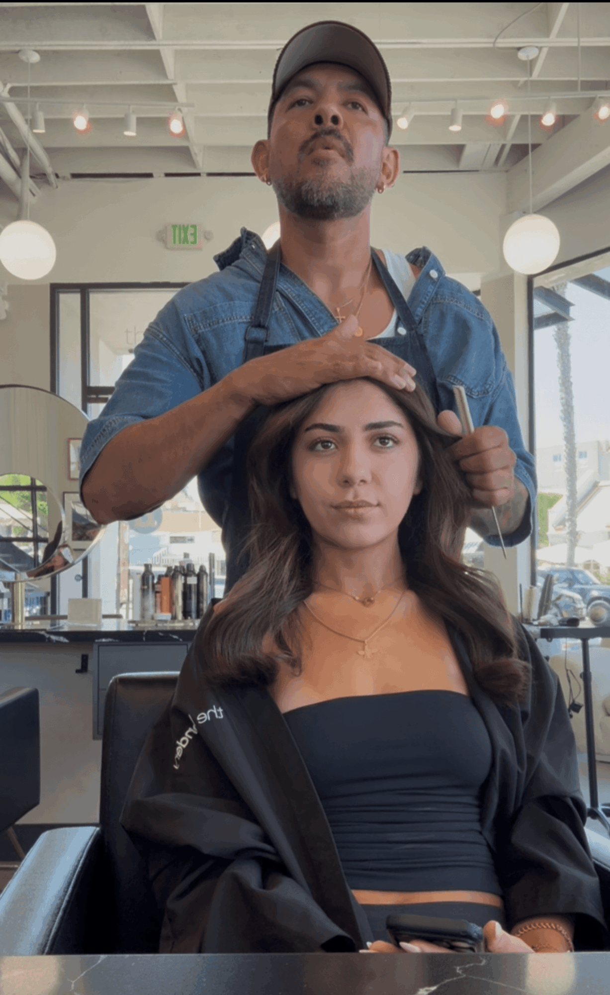 A woman with long dark hair sitting in a salon chair, a man standing behind her styling her hair, salon interior visible.