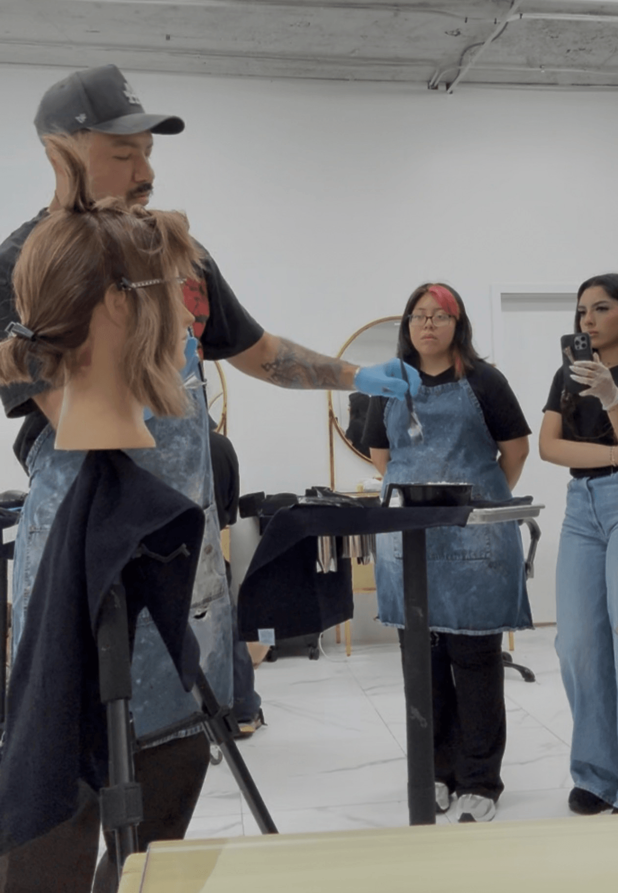 Instructor demonstrating hair cutting techniques to students in a classroom, with hair mannequins and tools on a table.