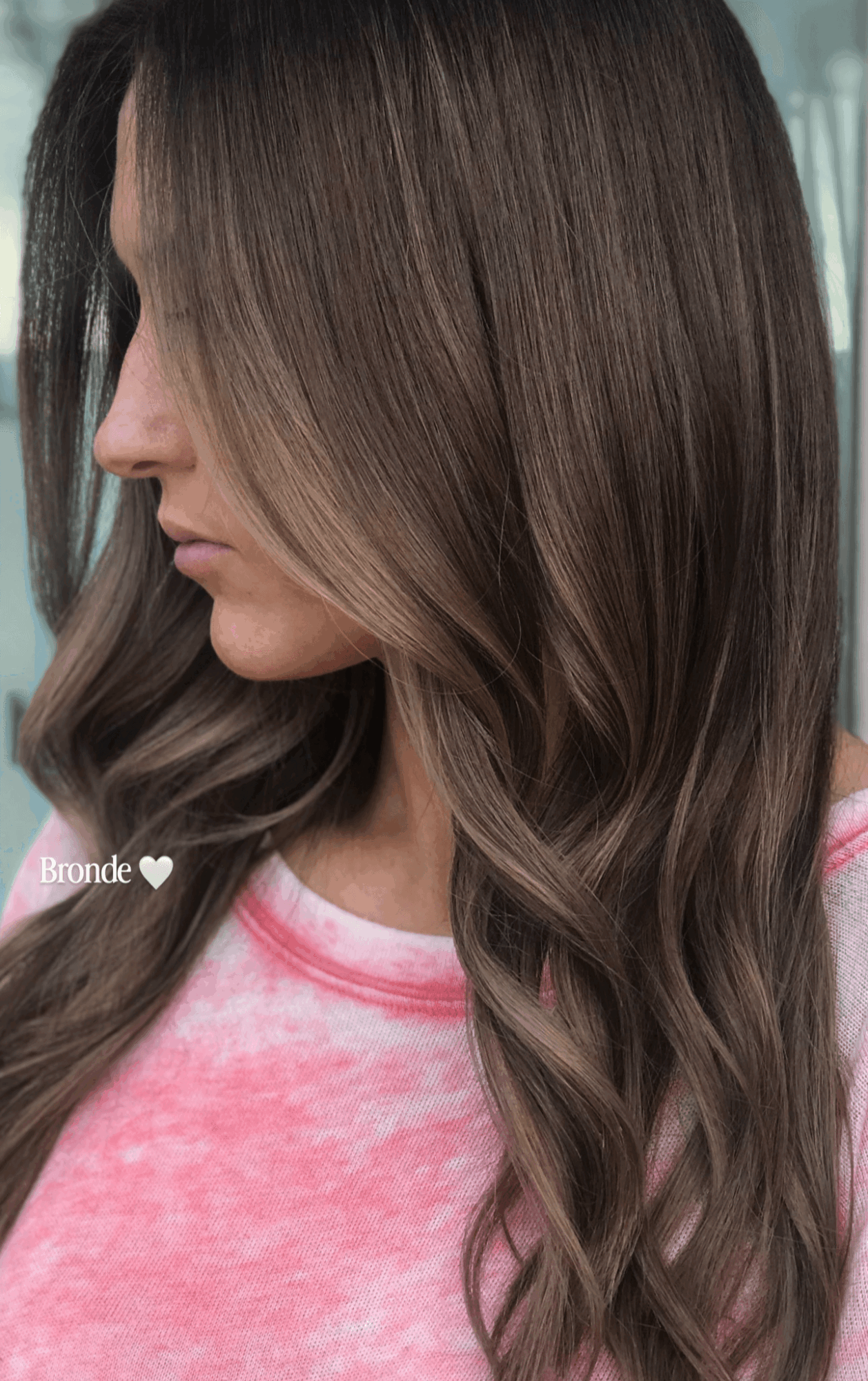 Side view of a woman with long, wavy hair with highlights, wearing a pink and white shirt.