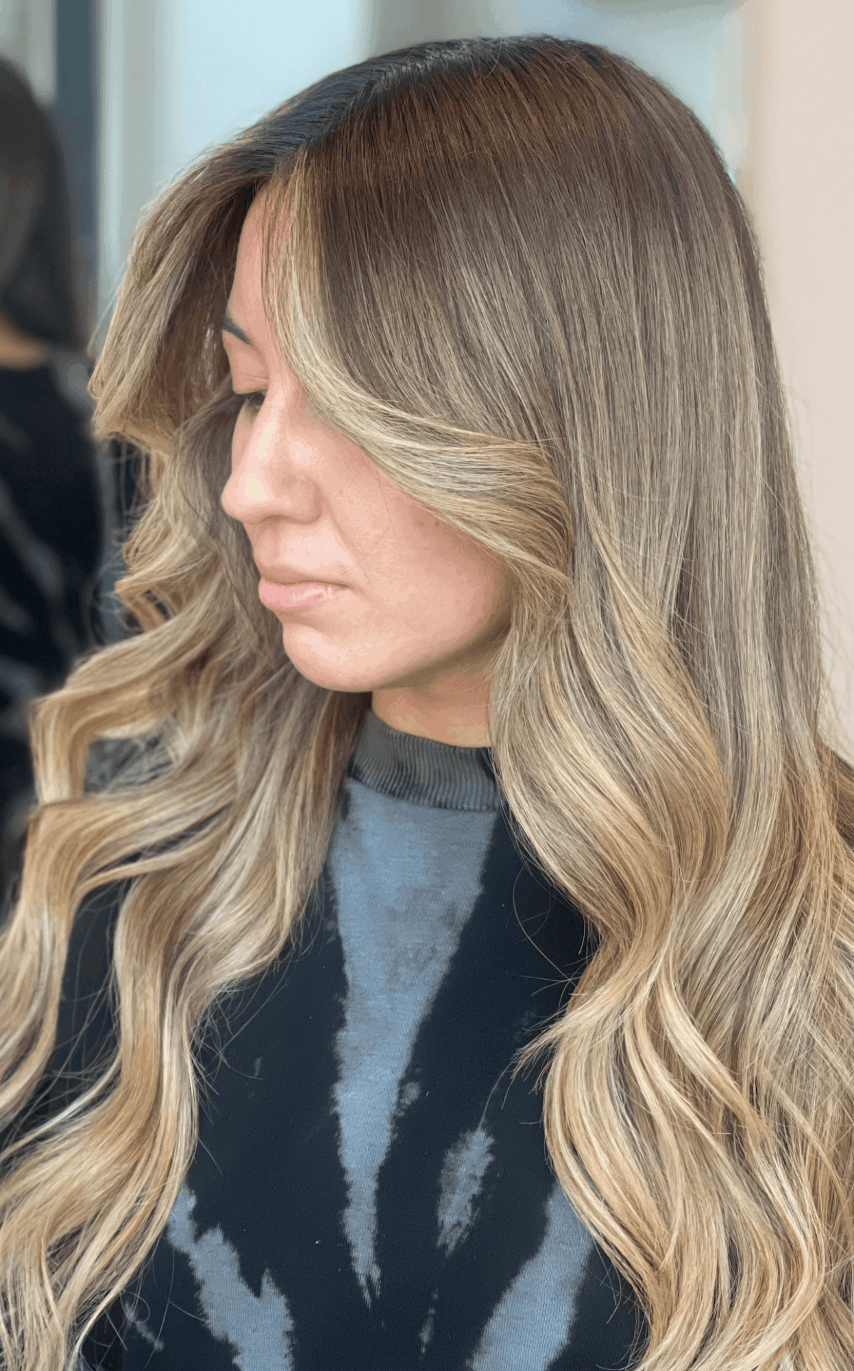 Side view of a woman with long, wavy, blonde hair and darker roots, wearing a dark top.
