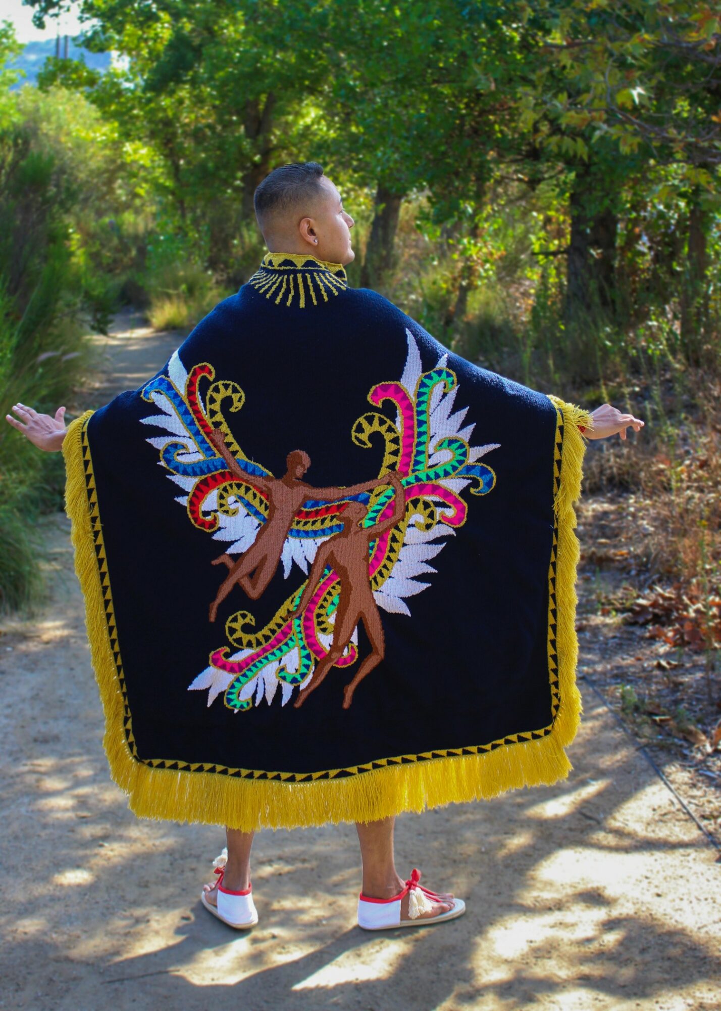 Person standing outdoors with arms outstretched, wearing a colorful, patterned shawl with a yellow border, trees in background.